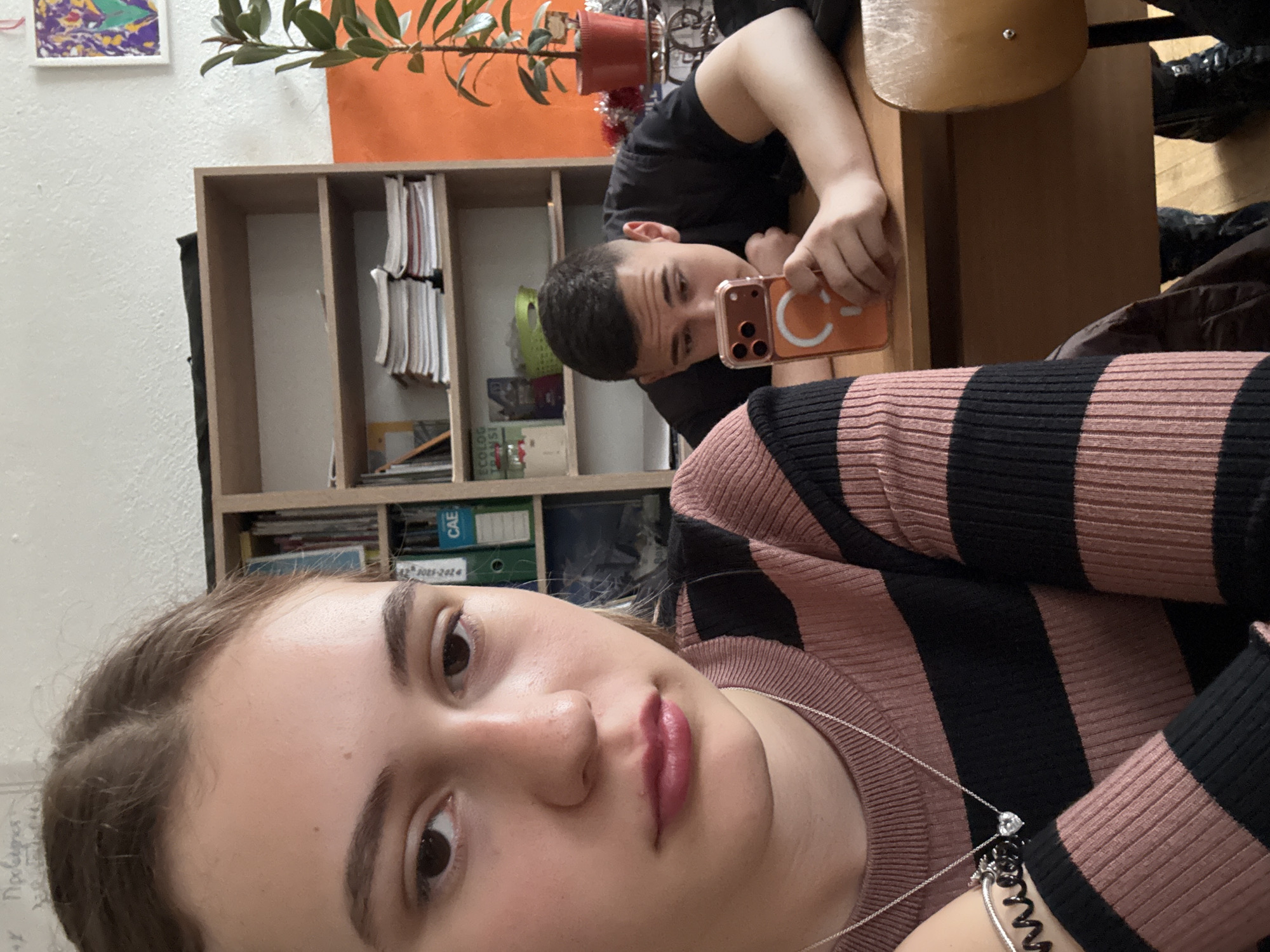 This indoor selfie, taken in Panagyurishte, Bulgaria, features two young individuals. In the foreground, a young woman takes the photo, looking directly at the camera with a neutral expression. She wears a pink and black striped long-sleeve top and a silver necklace with a heart pendant. On her wrist, a black spiral hair tie or bracelet is visible.
In the background, a young man with short hair is leaning on a wooden desk. He looks towards the camera, holding an orange smartphone with a distinctive white letter "C" on its case. Behind him, a wooden shelving unit is filled with books, folders, and papers. A green folder clearly displays the text "CAE" and "2021-2024". A potted plant sits on top of the shelves, with an orange wall section behind it. A small, colorful abstract artwork is framed on an adjacent white wall. The setting suggests a casual environment, possibly a classroom, study room, or home office. Faint Cyrillic text, possibly "Пробелы", can be seen on the white wall to the left of the woman's head. The overall mood is relaxed.
