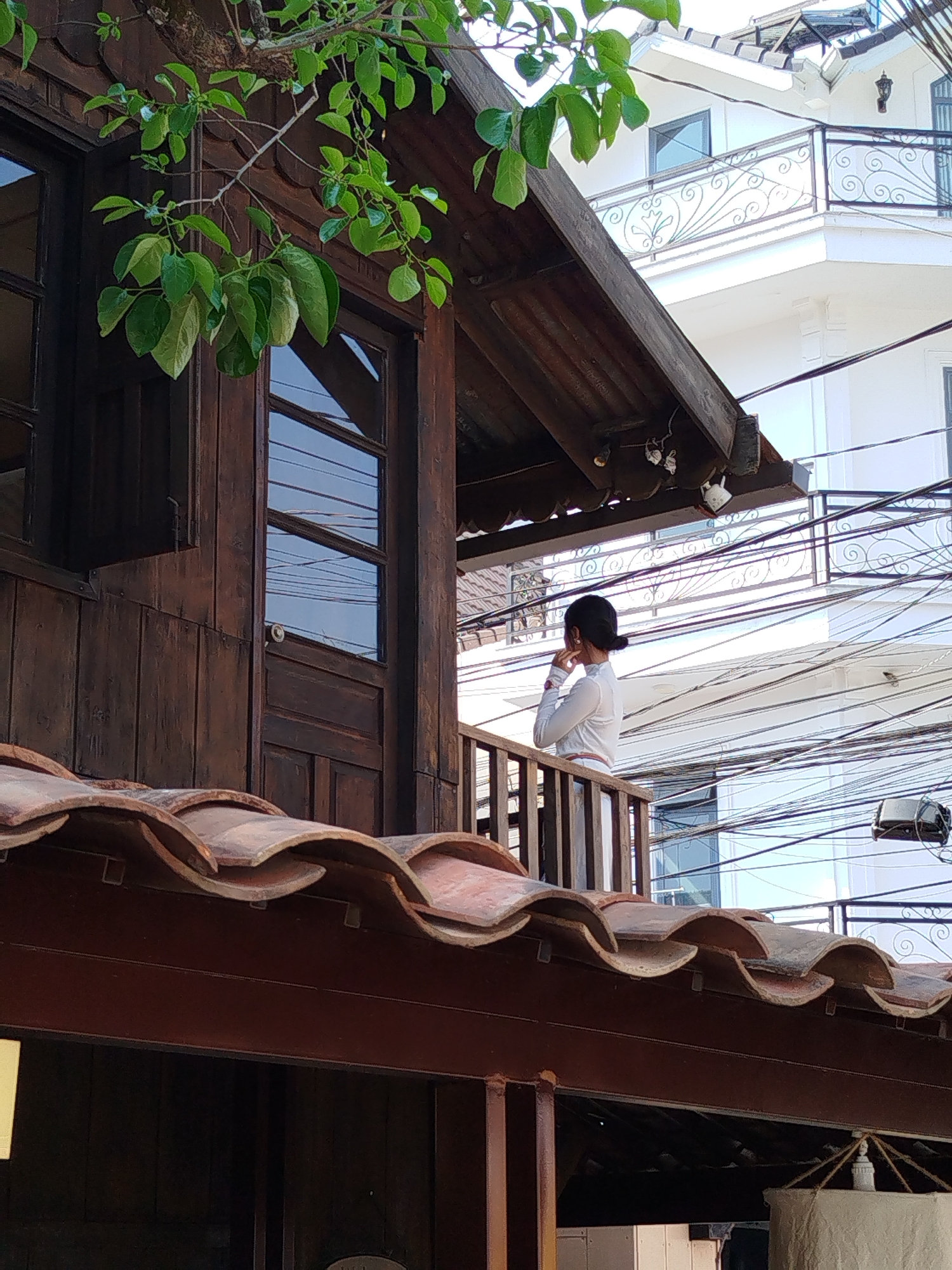 The image depicts a woman standing on a balcony in Da Lat, Vietnam. The woman is wearing a white long-sleeved shirt and has her hair pulled back in a bun. She is standing with her hand to her ear, as if listening or talking on the phone. The balcony is attached to a wooden building with a tiled roof. The background consists of other buildings and a tangle of overhead wires, typical of urban areas in Vietnam. The scene is partially framed by green foliage in the foreground.