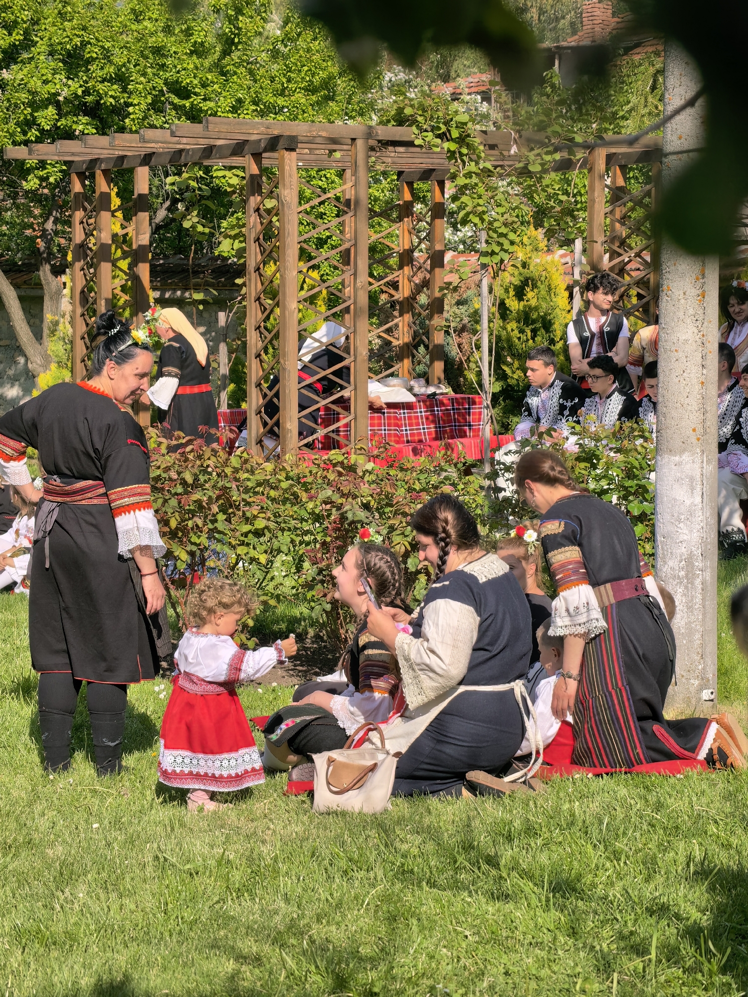 The image depicts a group of people in traditional Bulgarian clothing, gathered in a garden setting in the city of Sapareva Banya, Bulgaria. A wooden pergola is visible in the background, partially obscured by greenery. Several individuals are seated on a red blanket on the grass. One woman, dressed in black attire with white and red trim, stands next to a toddler wearing a red and white dress. The toddler is pointing towards the seated people. Other individuals are standing and sitting in the background, also dressed in traditional attire. The environment includes green grass, trees, and other foliage. The scene appears to be part of a cultural event or celebration.