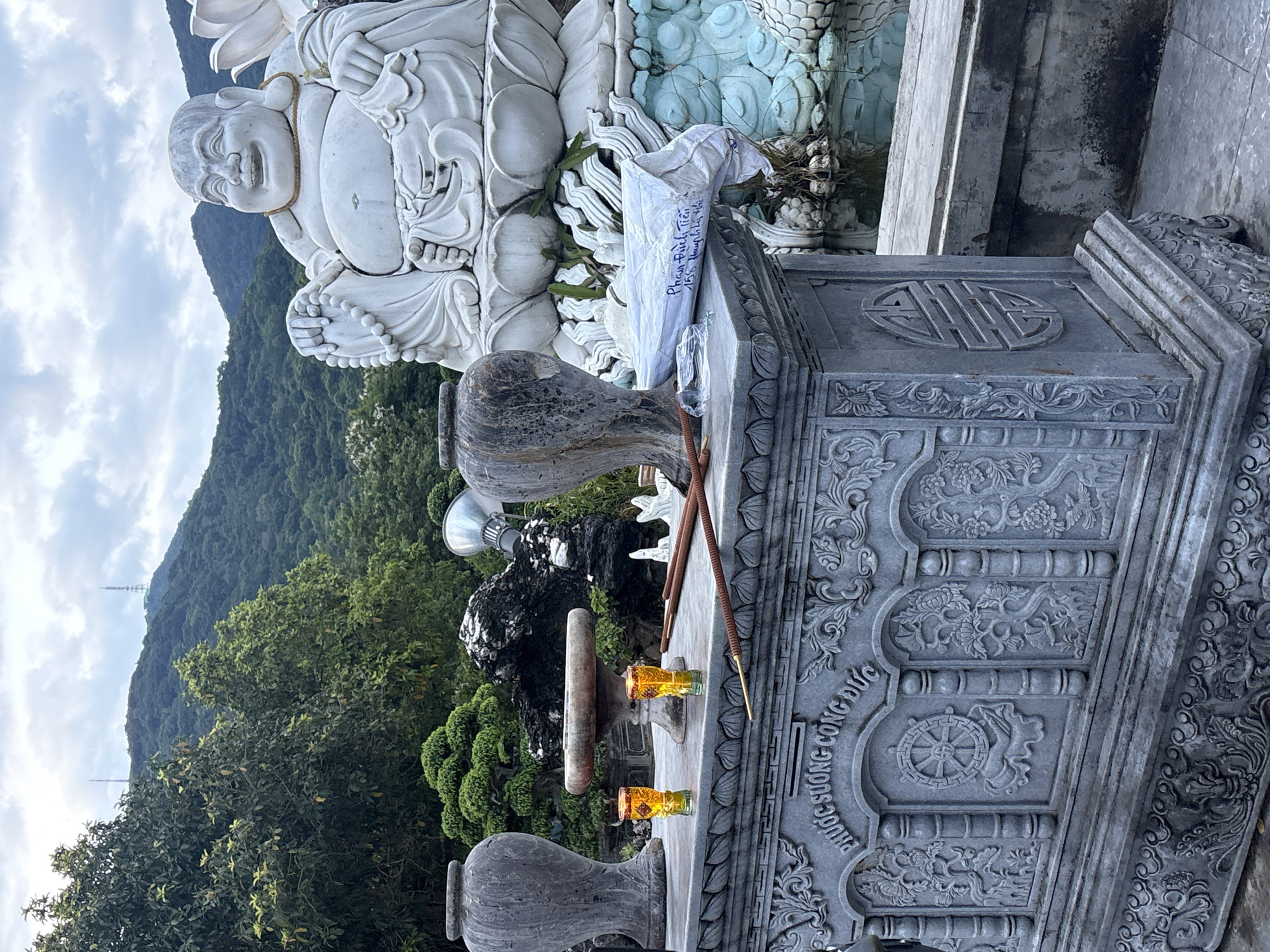 The image depicts an outdoor altar in front of a large stone statue of a laughing Buddha, likely located at a temple or religious site in Đà Nẵng, Vietnam. The altar is made of intricately carved gray stone, featuring floral motifs and what appear to be Buddhist symbols, including a Dharma wheel on the front panel. On the altar's surface are two small glasses containing a yellow liquid, possibly offering drinks, and several incense sticks, some of which are lit, emitting a slight haze. Two large stone vases flank the altar, and behind them are more decorative stone elements and lush greenery. In the background, a hillside covered in trees rises steeply, with a communication tower visible on its summit under a cloudy sky. The laughing Buddha statue is massive, seated on a lotus flower base, and appears to be made of white stone. The scene suggests a place of worship or remembrance, possibly connected to the Buddhist faith, a significant religion in Đà Nẵng.
