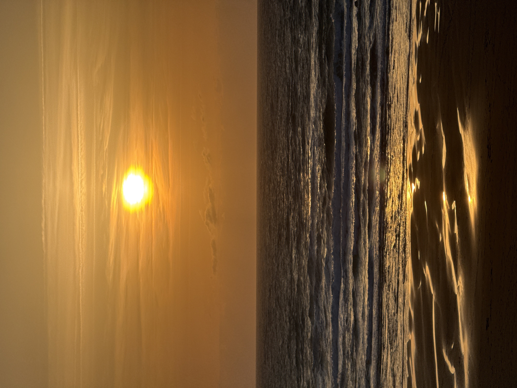 The image captures a serene sunset over the ocean at Sopela, Spain. The sun, a bright orb of light, is positioned in the upper center of the frame, casting a warm, golden glow across the sky and reflecting on the water. The sky is a gradient of oranges and yellows, with wispy clouds streaking horizontally. The ocean surface is textured with waves, some breaking into white foam closer to the shore. The foreground reveals a sandy beach, wet from the receding tide, with shallow pools of water creating mirror-like reflections of the sunset. The overall scene is peaceful and evokes a sense of natural beauty.