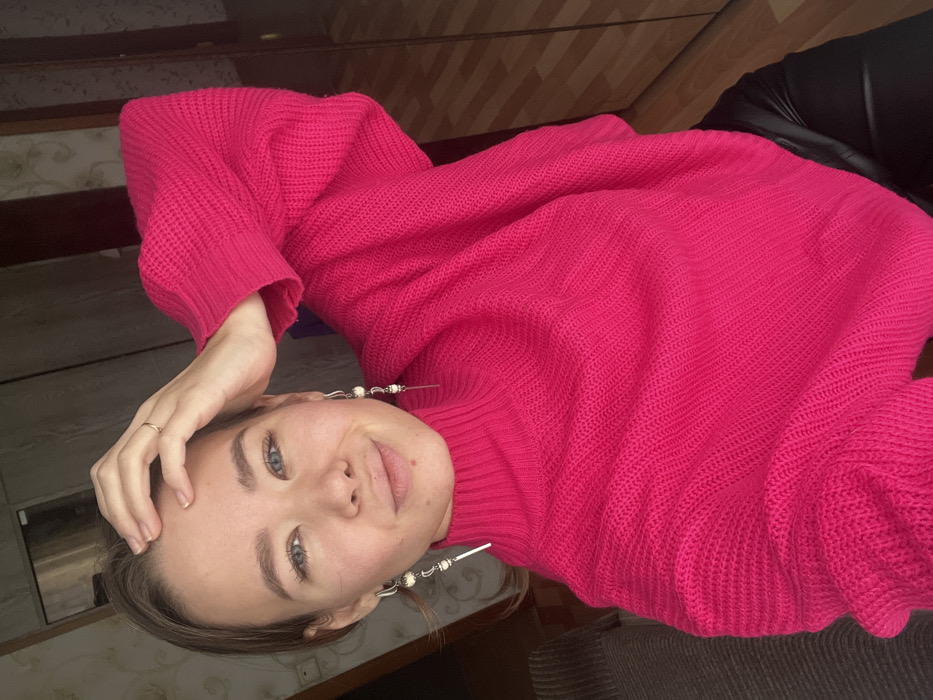 A woman is photographed from a tilted perspective, wearing a bright pink knitted sweater and dark leather pants. She has fair skin, blue eyes, and her hand is near her forehead. She is wearing silver earrings with white beads. The background includes wooden panels and a beige patterned wallpaper.