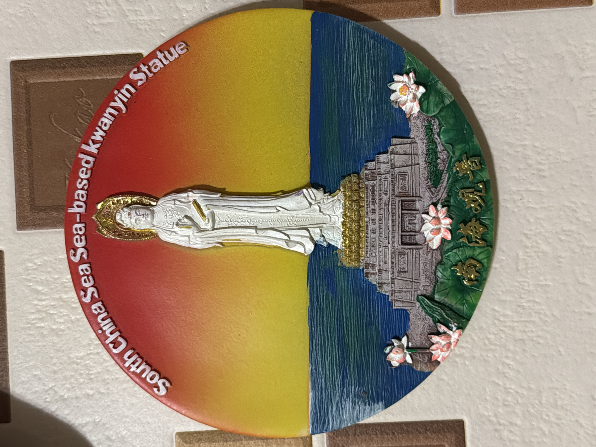 The image depicts a circular souvenir featuring a representation of the South China Sea Sea-based Kwanyin Statue. The souvenir is divided vertically into two halves. The left half features a gradient of colors ranging from red at the bottom to yellow at the top, with the text "South China Sea Sea-based Kwanyin Statue" around the edge. The right half shows a depiction of the sea in blue and a temple structure with lotus flowers around it. A white statue lies across the diameter of the circle. The background appears to be a tiled wall. The image does not have a connection to Krasnoyarsk, Russia.