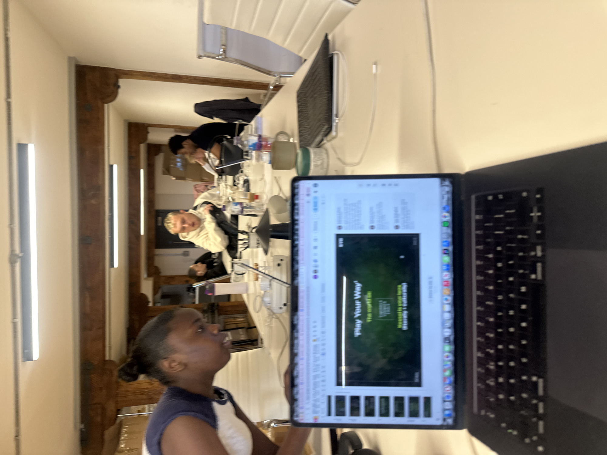 The image shows a group of people gathered around a long table in what appears to be a modern office or co-working space. The location is suggested to be in the city of London, United Kingdom.
**People:** Several individuals are visible, some closer to the camera than others. One woman is in the foreground, looking towards the table and a laptop in front of her. Other individuals are seated further down the table, some of whom are looking down at their own devices or papers.
**Objects:** A long table dominates the center of the scene, upon which various items are placed, including a computer monitor, mugs, and water bottles. A laptop is prominently displayed in the foreground with a webpage displaying the words "Play Your Way."
**Environment:** The setting appears to be a well-lit, modern office space. There are white walls and wooden beams. Vertical light bars are on the wall, and there are chairs, a modern keyboard, and office equipment present.