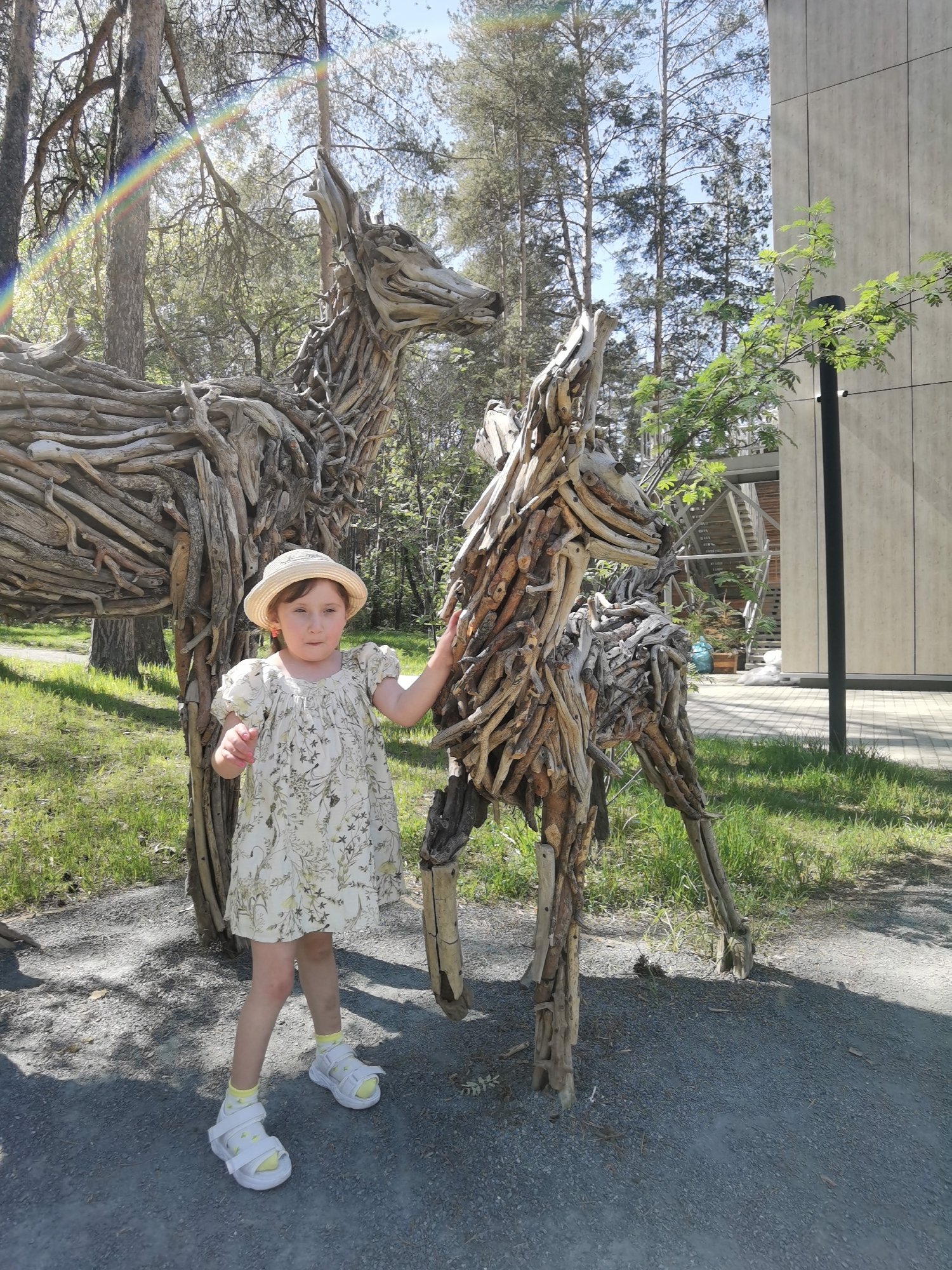 A daytime shot in Perm, Russia, shows a young child in a straw hat and patterned dress standing next to two wooden sculptures of horses. The child is fair-skinned and has her right hand on the sculpture's neck. The sculptures are made of sticks and logs. There are trees and a building in the background. The ground is covered in gravel. A rainbow is visible in the upper left corner of the frame.