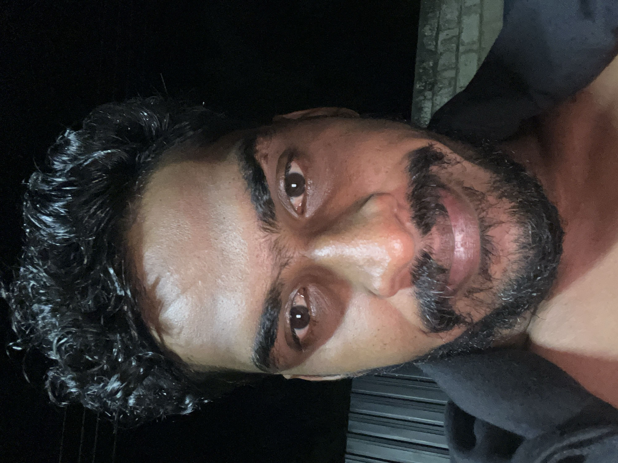 A close-up portrait shows a man's face, tilted 90 degrees to the right, against a dark, indistinct background. The image was taken at night in Arugam Bay, Sri Lanka. He has a medium to dark complexion, with dark, curly hair that appears wet or styled, catching highlights from an unseen light source. He sports a well-maintained dark beard and mustache. His dark brown eyes are open wide and look directly at the viewer, conveying an intense, perhaps playful or challenging expression. His eyebrows are slightly furrowed, and a subtle smirk is visible on his lips. Harsh lighting illuminates his face unevenly, highlighting his forehead, nose, and cheekbones, and creating a sheen on his skin that could be sweat or oil. A dark-colored garment, possibly a hoodie or shirt, is visible around his neck and shoulder area. In the upper right background, faint vertical lines suggest a distant structure or wall. There is no visible text.