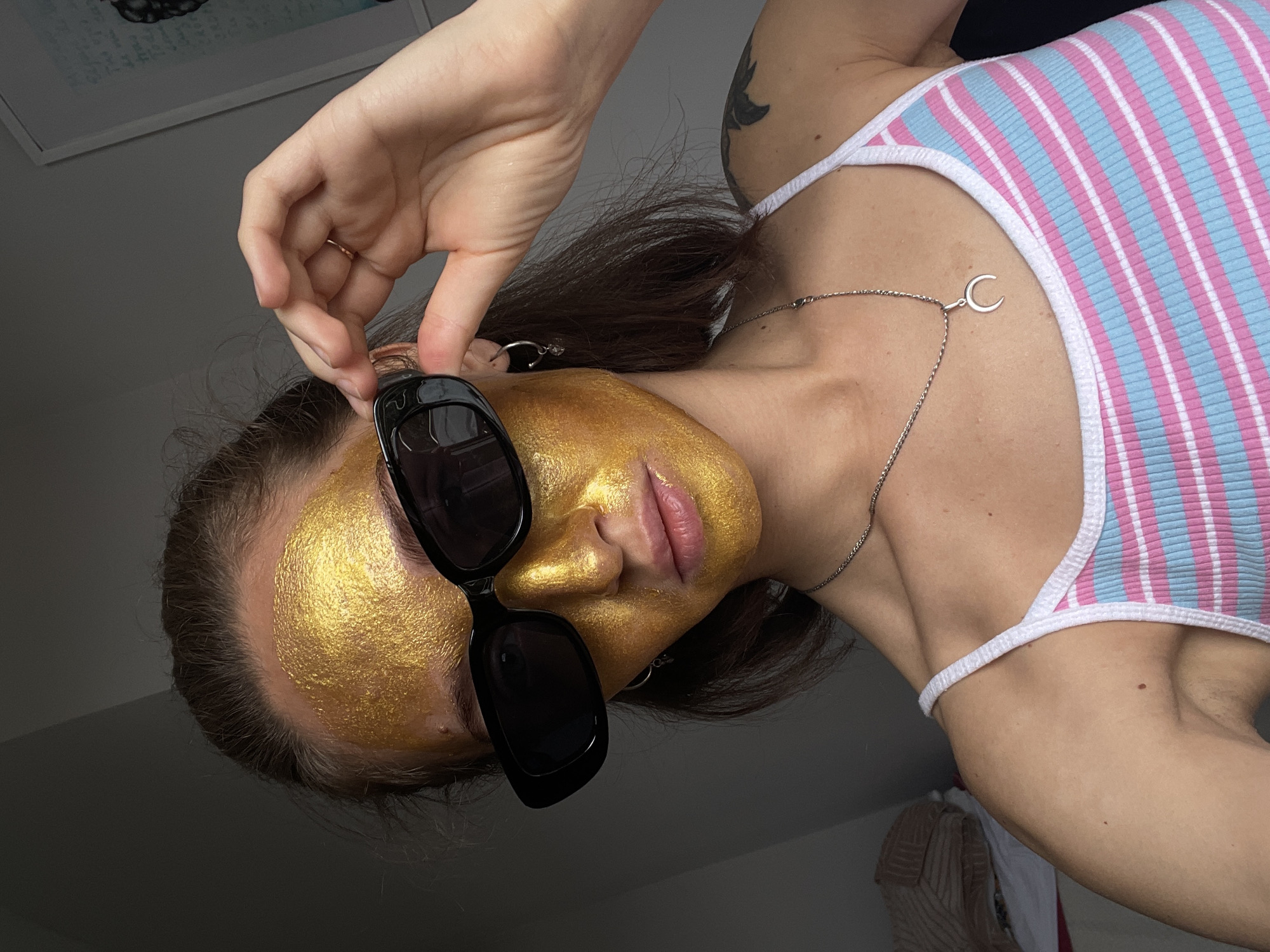 A woman with a gold facial mask and black sunglasses is depicted indoors. She wears a striped pink and blue tank top with a white trim. A silver necklace with a crescent moon pendant hangs around her neck. Her right hand is holding the frame of her sunglasses. There is a tattoo on her upper arm. The woman has dark hair. The background is blurry. The image is taken from a low angle and is slightly tilted. There is no evidence indicating that the photo was taken in Sochi, Russia.