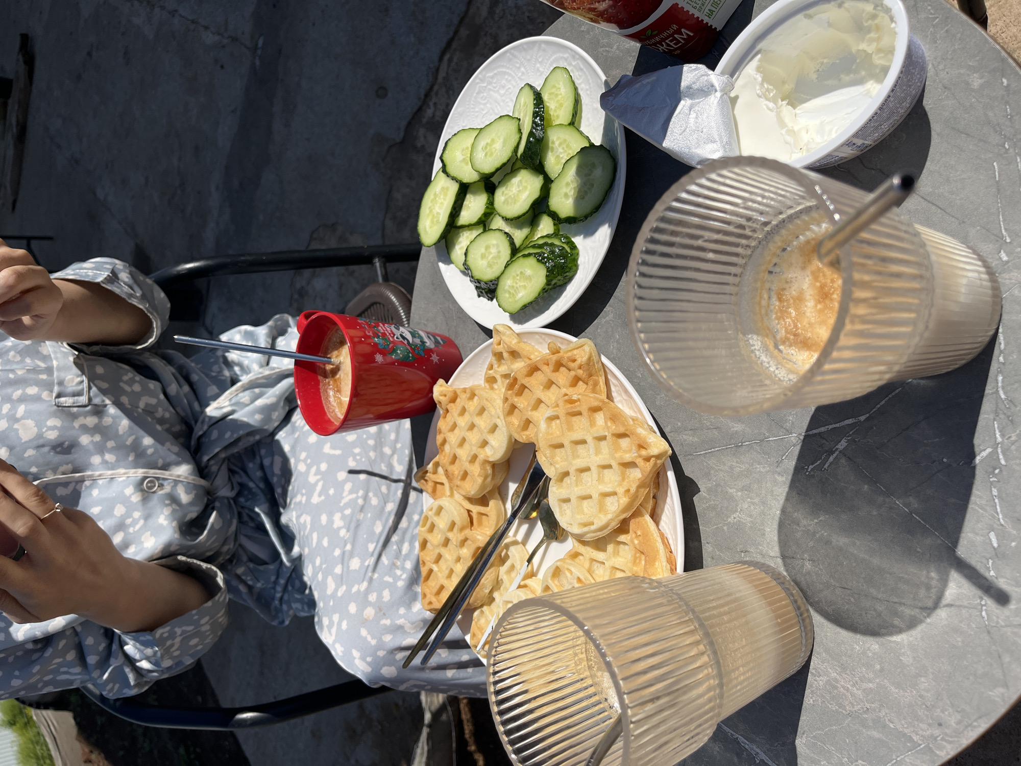 The scene shows a person wearing blue patterned pajamas sitting at a gray marble-look table, possibly outdoors. On the table are several food items: a plate of sliced cucumbers, a container of white cream cheese, a plate of heart-shaped waffles with two forks, a red mug, and two tall glasses with straws, one containing a coffee drink. The scene suggests a breakfast setting. The location is Лобаново, Russia.