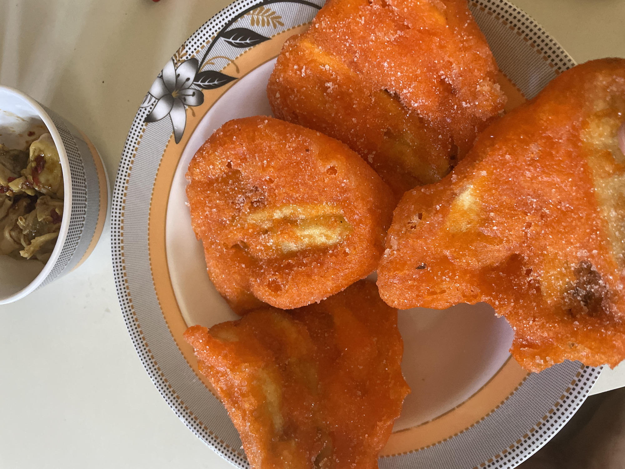 The image shows a plate of turon, a Filipino snack made of sliced bananas coated in brown sugar, wrapped in spring roll wrapper, and deep-fried. The turon pieces are orange and appear to be coated with granulated sugar. The plate has a floral pattern in white, black, and gold. Next to the plate is a cup filled with an unidentified food item. The location is Davao City, Philippines.