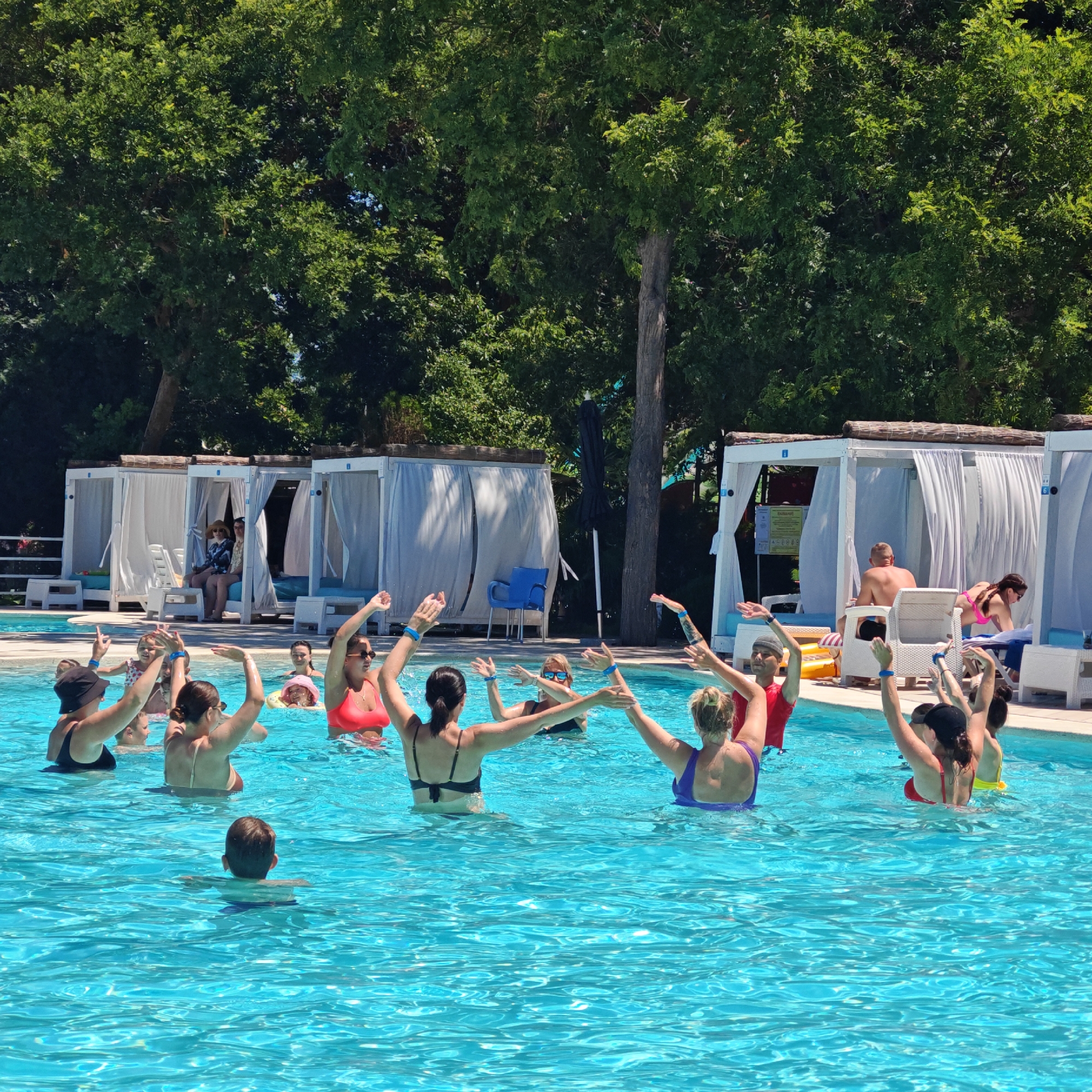 In the city of Shchelkino, Ukraine, a group of people are participating in a water aerobics class in an outdoor swimming pool. The water is a vibrant turquoise, reflecting the clear blue sky above. Several individuals, mostly women, are submerged in the water up to their waists and chests, their arms raised and extended in various poses characteristic of an exercise routine. They are wearing swimwear in a range of colors, including red, purple, and black. Some participants have blue wristbands on. In the background, there are white cabanas with draped curtains, suggesting a resort or recreational facility. Behind the cabanas, lush green trees form a dense backdrop. A few other people are visible in the distance, some seated and observing the activity in the pool, including a man and woman in one cabana. A young boy is also in the pool, further back and facing away from the camera. The overall scene depicts a lively and active summer day at a poolside location.