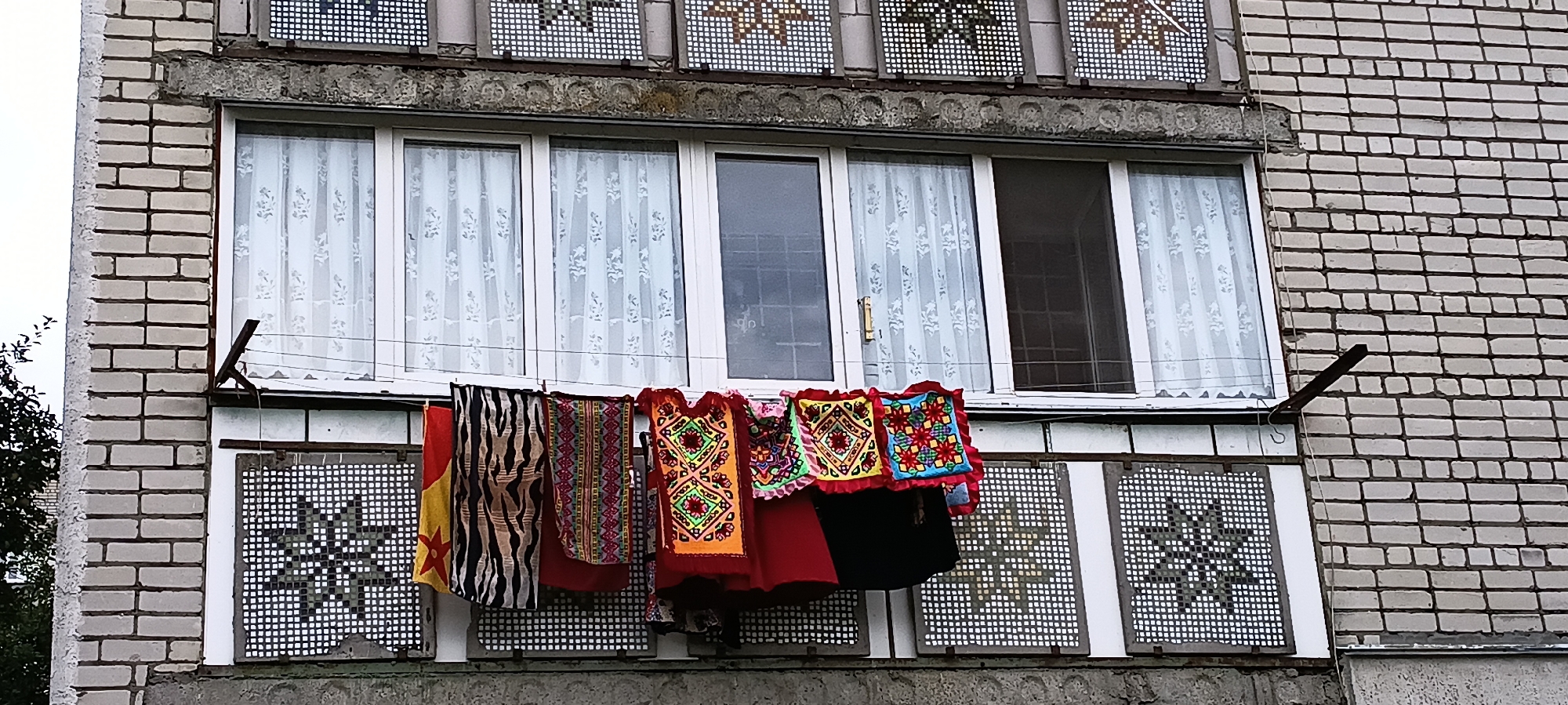 The image shows a close-up view of a balcony on a brick building in Kamianka, Ukraine. The balcony has white window frames with white, patterned curtains visible behind them. Below the windows, several colorful, embroidered garments are hanging on a clothesline. These include what appear to be vests or tops with geometric patterns and bright colors like orange, red, green, and blue, as well as a striped fabric and a piece with a zebra-like pattern. The garments are attached to a wire clothesline supported by metal brackets extending from the building. Below the clothesline are decorative panels with a white grid pattern and a star-shaped motif. The brickwork of the building is visible on the right and left sides of the image, and the sky is overcast.