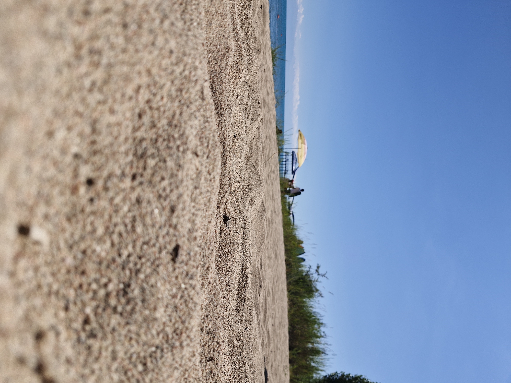 The image depicts a low-angle, close-up view of sandy beach terrain in Bulan-Segettu, Kyrgyzstan, on a clear, sunny day. The foreground is dominated by the granular texture of the sand, with a sharp focus on the individual grains and small debris. In the mid-ground, a body of water, likely Lake Issyk-Kul, stretches across the horizon under a bright blue sky. A few distant figures are visible near a pier, some shaded by a yellow and blue umbrella. Green vegetation and tall grass line the edge of the beach behind the sand. The overall scene suggests a tranquil summer day at a lakeside resort.