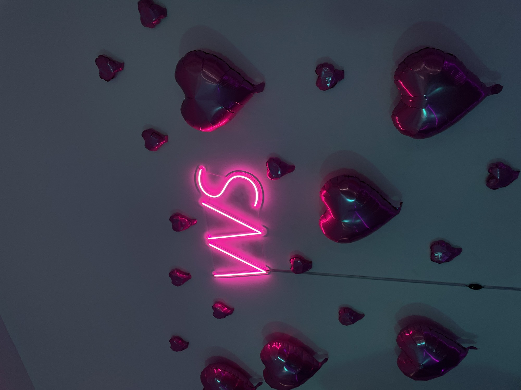The media file captures an indoor decorative display featuring numerous heart-shaped foil balloons and a prominent neon sign against a dark, plain wall or ceiling.
The central subject is a bright pink neon sign spelling the letters "VS". The vibrant glow of the neon illuminates the immediate surroundings, casting a strong magenta hue and reflections onto the nearby surfaces and objects. A thin, dark wire extends horizontally from the right side of the neon sign.
Scattered across the frame are many heart-shaped foil balloons. These balloons are predominantly pink or magenta, reflecting the light from the neon sign. Their metallic surfaces also display cool blue and cyan reflections, particularly in the shadowed areas and highlights, indicating their glossy texture. The balloons vary in size and are arranged in a seemingly random pattern, creating a festive or romantic atmosphere. Shadows cast by the balloons suggest they are affixed to the surface, protruding slightly.
The overall lighting is dim, emphasizing the luminous neon sign and the reflective quality of the balloons. No people or specific events are depicted; the image is a static shot of interior decoration. The arrangement and colors evoke feelings of love, celebration, or a modern, stylish aesthetic.
While the location context provided is Saint Petersburg, Russia, there are no discernible visual cues within the image itself to identify this specific geographical location.