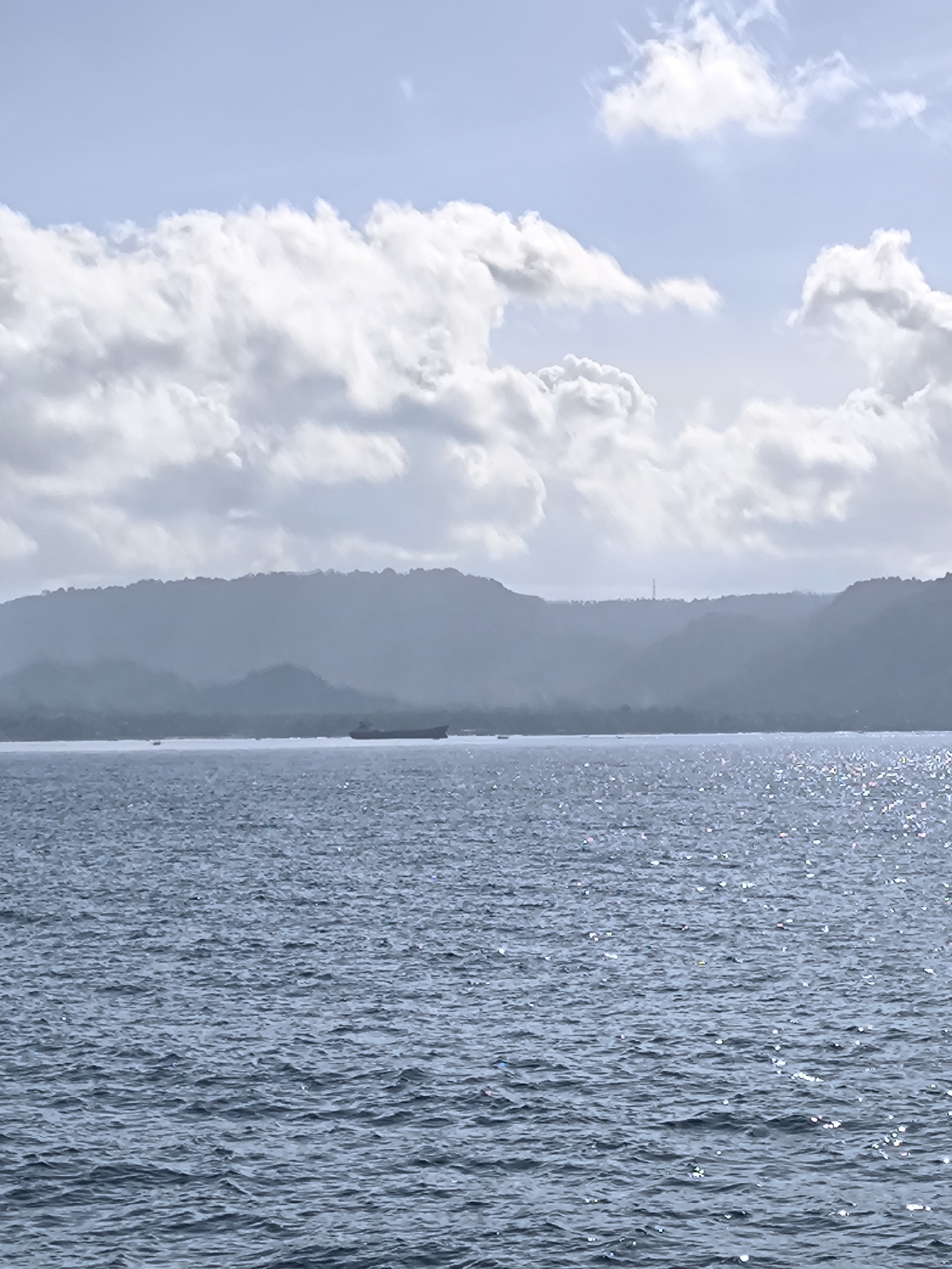 The image depicts a body of water, likely the sea, with gentle ripples and reflective glints of sunlight scattered across its surface. In the distance, a hazy silhouette of land is visible, possibly hills or mountains, with a darker mass resembling a large ship positioned near the horizon. Above the land, a sky filled with white and gray clouds fills a significant portion of the upper frame. The location is likely in Unknown, Indonesia, based on prior instruction.