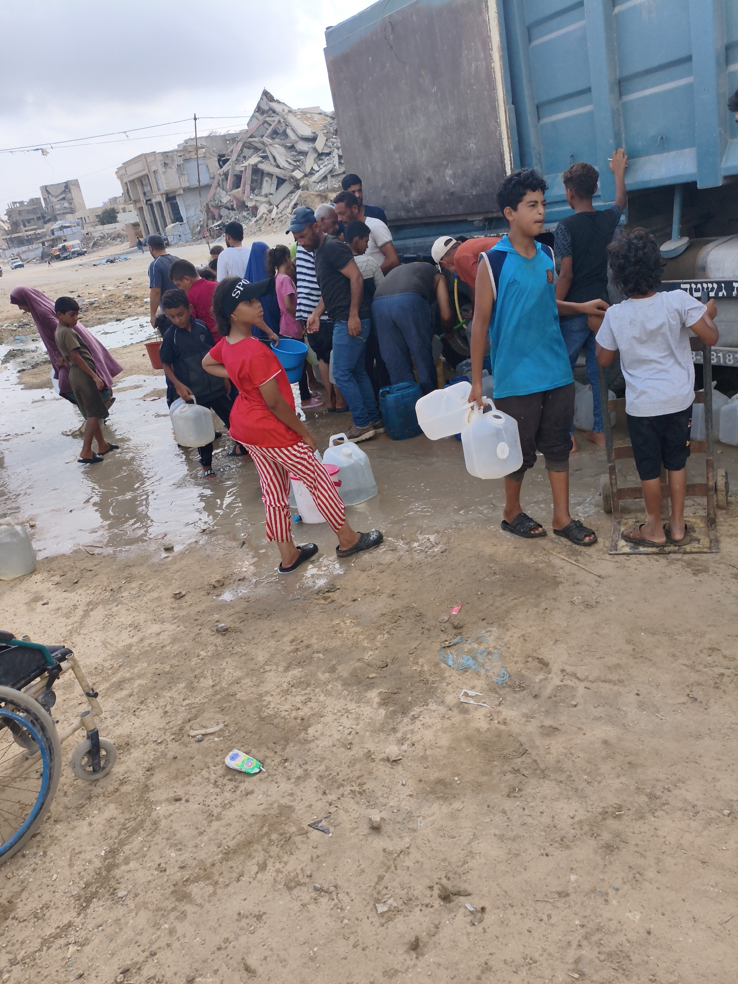 In an outdoor scene of destruction in an unknown city, a group of people, including men, women, and children, are gathered around a blue truck, collecting water in plastic containers. The ground is muddy and littered with debris, and destroyed buildings are visible in the background. A wheelchair is also present in the foreground. The sky is overcast.