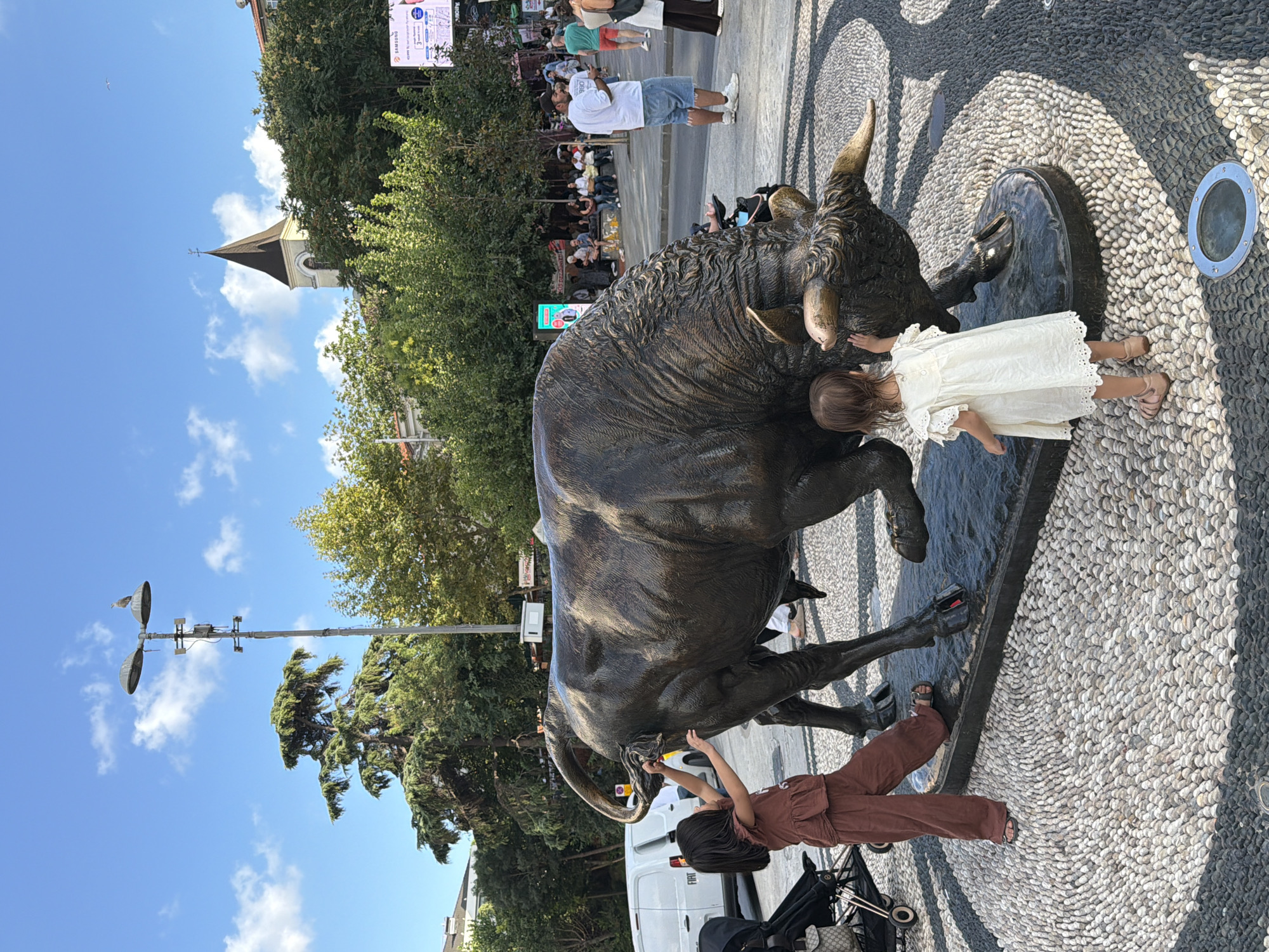 A bronze sculpture of a bull stands in a public square in Kadıköy, Turkey. Two young girls are interacting with the sculpture; one is touching its tail, and the other is touching its flank. The ground is paved with pebbles in a mosaic pattern. In the background, there are trees, a street lamp with a bird perched on it, a building with a steeple, and several people walking or sitting. A white van is parked to the left of the sculpture. The sky is blue with scattered clouds.