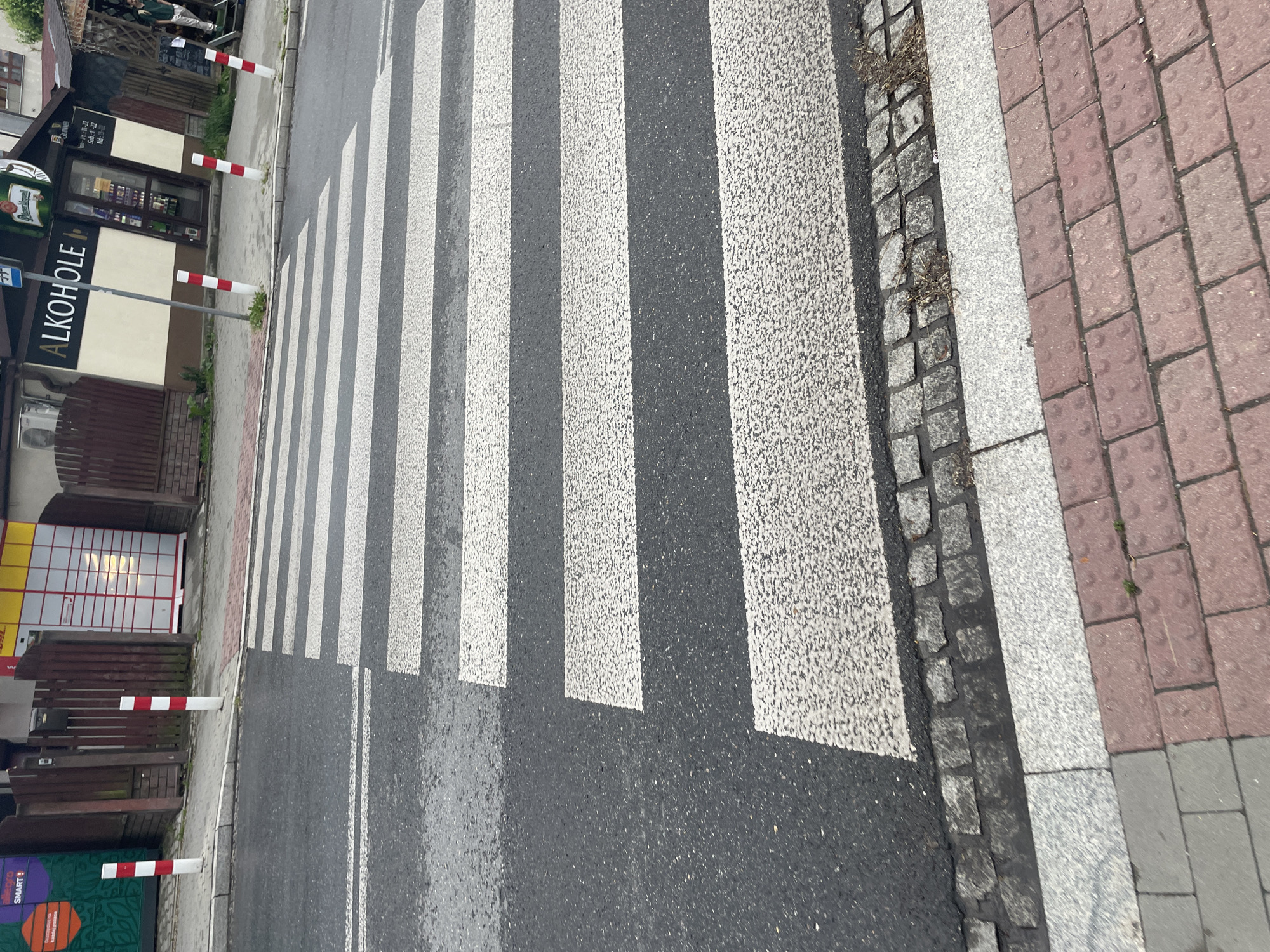 The image presents a street scene dominated by a pedestrian crossing under bright daylight. The asphalt road features prominent white zebra stripes. To the right of the crossing, the sidewalk is composed of reddish-brown tactile paving with raised bumps, transitioning to light grey paving stones, and edged by small, dark grey cobblestones.
On the opposite side of the street, a parallel sidewalk is delineated by several white and red striped bollards. Behind these bollards, a commercial building features a storefront. A large sign with the word "ALKOHOLE" (Polish for alcohols) is prominently displayed on its facade. Above this, a round sign partially shows the words "PIWO" (beer) and "GUINESS." Further down the street, two automated parcel lockers are visible: a green and orange one bearing the "SMART!" logo, and a yellow and red DHL locker.
In the background, near the "ALKOHOLE" store, two individuals are partially visible. The scene appears to be static, with no discernible activity or movement. The bright, clear lighting suggests it is daytime, likely a sunny day. The visible Polish text "ALKOHOLE" and "PIWO" indicates a location in Poland or a Polish-speaking region. The image captures an everyday urban environment.