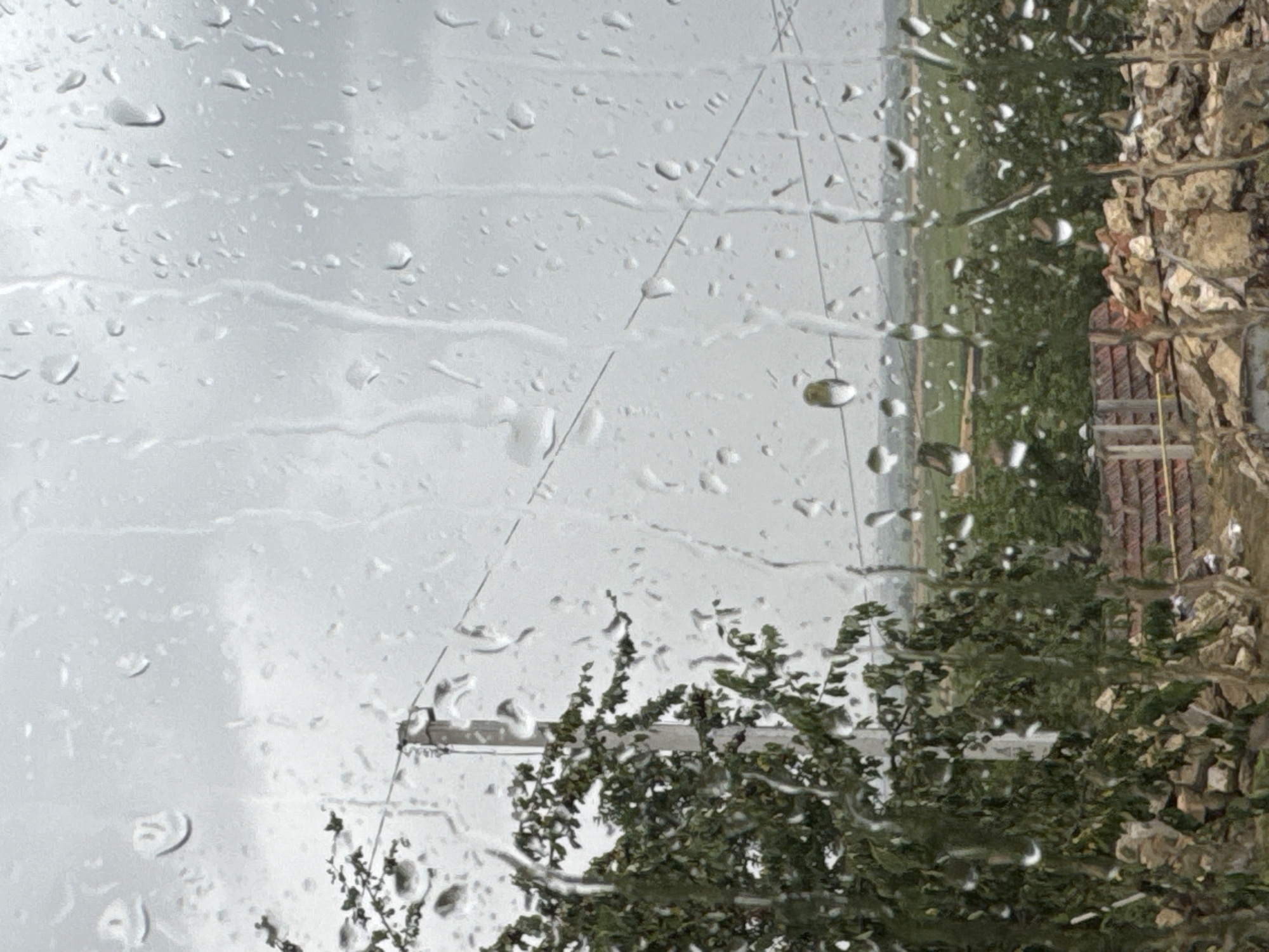 This is a photograph taken through a window covered in raindrops. The scene outside is a blur of greenery and what appears to be a stone wall or building, suggesting an outdoor setting. The primary focus is on the water droplets themselves, which distort and refract the view beyond, creating an abstract and textured effect. The raindrops vary in size and shape, with some appearing as perfect spheres and others elongated or streaked, indicating movement or the force of the rain.
The background, though obscured, hints at a landscape with trees and possibly a field or open space. A thin, dark line runs diagonally across the frame, possibly a wire or string. On the right side, a textured surface, likely stone, is visible, along with hints of red tile, suggesting part of a building. The overall impression is one of a rainy day, with the moisture on the glass creating a diffused and softened image of the outside world. There is no visible text in the image. The lighting suggests it is daytime, but the overcast conditions due to the rain prevent clear identification of the time of day. The mood is somewhat melancholic, typical of rainy weather, with the raindrops creating a sense of separation between the viewer and the external environment.