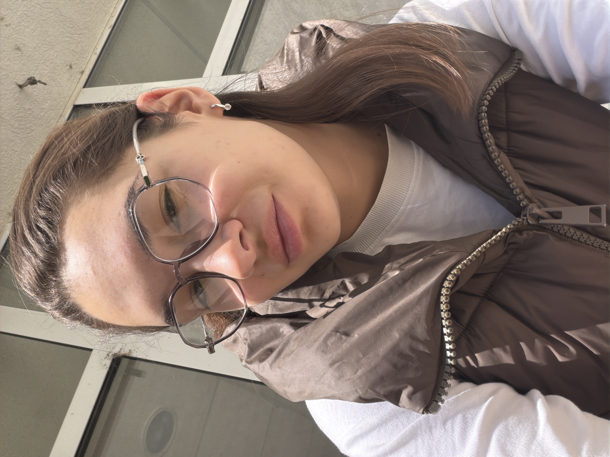 A close-up, sideways-oriented portrait captures a person with long, straight dark brown hair, looking downwards towards the camera with a neutral to slightly serene expression. They wear clear-framed glasses and a small silver hoop earring with a clear stone on their upper left ear. The person is dressed in a white t-shirt with a ribbed collar and a brown, shiny puffer vest featuring a prominent metallic zipper with large teeth.
The image is taken outdoors in bright, direct sunlight, evident from the distinct shadows cast on the clothing and facial features, suggesting a sunny day. The background consists of a light-colored, possibly textured wall or building exterior with reflections from window panes or glass surfaces. A small, dark, irregular object, possibly debris, is visible on the wall behind the person's head. The overall setting implies an urban environment, specifically Sofia, Bulgaria. There is no visible text in the image.