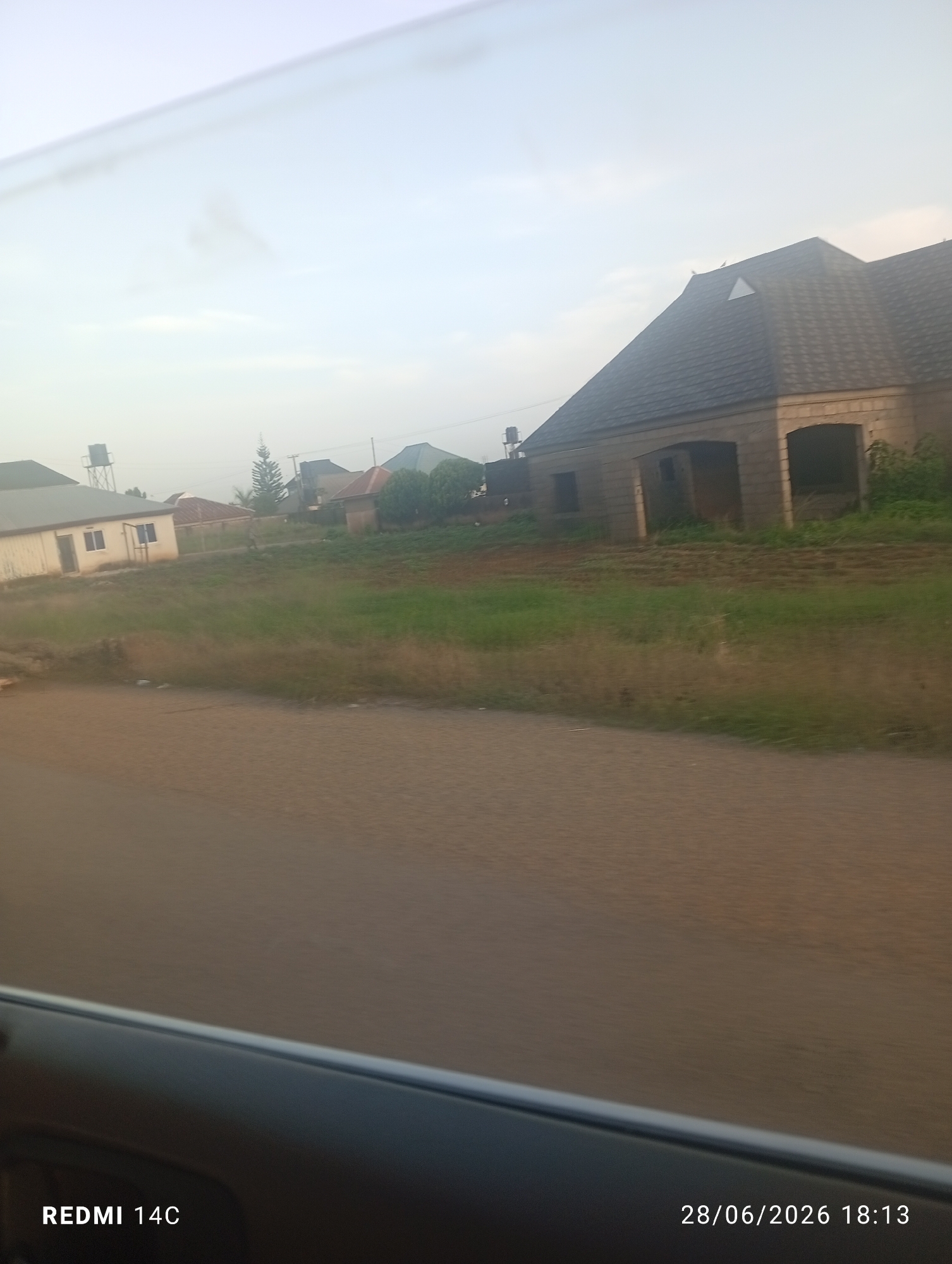 The image captures a residential area in Jos, Nigeria, possibly during overcast conditions. In the foreground, a paved road occupies the lower portion of the frame, with a dark, reflective car interior edge visible at the bottom. The midground reveals a grassy slope dotted with several buildings under construction or renovation. One prominent structure in the right midground features a dark, multi-pitched roof and a light-colored brick exterior with large arched openings. To the left, a simpler, light-colored building with a flat roof and a water tank on a tower is visible. The background shows more houses, some with tiled roofs, and sparse vegetation, including a tall conifer tree. The sky is a pale, washed-out blue, suggesting diffused light. The timestamp "28/06/2026 18:13" and "REDMI 14C" are present in the bottom right and left corners, respectively. There are no people visible in the scene.