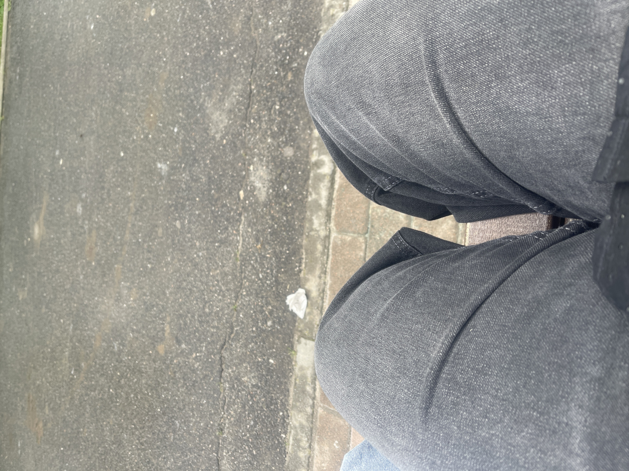 The image presents a downward, first-person view of a person's legs and the ground beside them. The person is wearing dark grey or black jeans, with folds and creases visible in the fabric over their bent knees. A small sliver of a lighter blue fabric, possibly another layer of clothing or the bottom of the jeans, is visible at the very bottom edge. The person appears to be sitting or standing with their legs bent, possibly on a bench or a low wall, as indicated by a narrow, wooden or brick-like surface directly beneath their thighs.
To the left of the legs is a ground surface, primarily composed of grey asphalt or pavement. This surface shows signs of wear, with visible cracks, small white and dark specks, and slight discolored patches that could be dirt or stains. Running parallel to the person's position and separating them from the main paved area is a strip of lighter-colored, possibly brick or stone pavers. A small, crumpled piece of white litter, possibly paper or plastic, rests on this lighter strip, near the person's lower leg. The lighting suggests it is daytime, likely overcast, as there are no strong shadows. No specific activity is depicted beyond the person's presence. There is no visible text in the image. The setting appears to be an outdoor urban or suburban environment in Poltava, Ukraine.