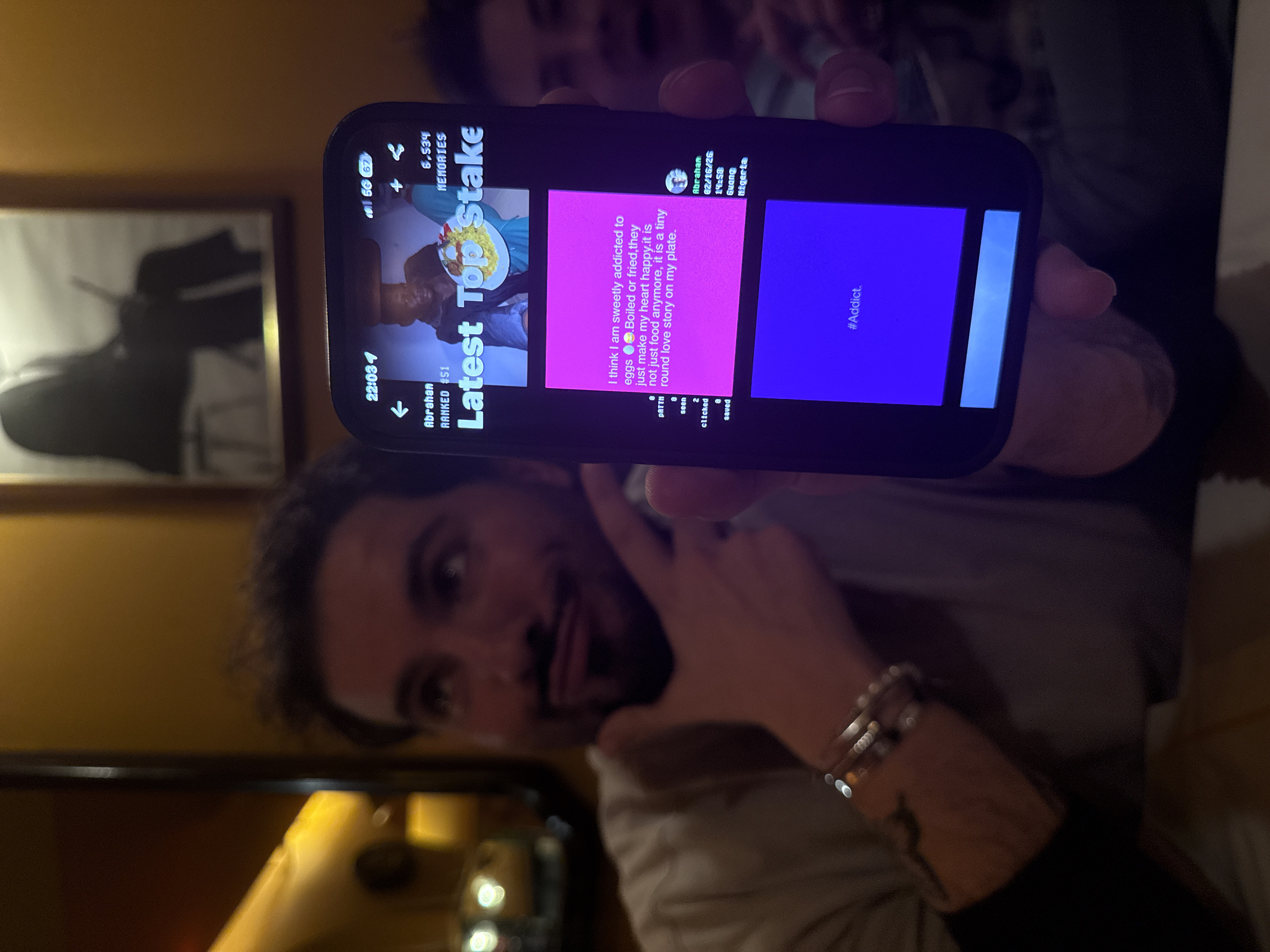 The image captures a casual, indoor scene, likely in the evening given the warm, artificial lighting. The primary subject is a person, appearing to be a man with dark hair and a beard or stubble, holding a smartphone up towards the camera. He is looking directly at the camera with a playful expression, sticking his tongue out slightly. He wears a light-colored long-sleeved shirt or top, and several metallic bracelets or bangles are visible on his left wrist, along with what appears to be a small tattoo on his forearm.
The smartphone, a dark-colored device resembling an iPhone, is held in portrait orientation but the image is rotated, making the screen appear sideways. The screen is brightly lit, displaying an application interface, possibly a social media platform or a personal journaling app.
The content on the phone screen includes:
- **Top Bar:** Time "22:03", a cellular signal icon, "5G" network indicator, and battery percentage "67".
- **Header Section:** A back arrow, the name "Abrahan", and "RANKED #51" are visible on the left. On the right, icons for sharing (paper airplane), adding content (+), and "6,534 MEMORIES" are displayed.
- **Main Content Area:** The phrase "Latest Top Stake" is prominent, appearing vertically and partially overlapping an image. This image shows a plate of food, possibly rice with other ingredients like eggs and vegetables, held by hands wearing a light blue sleeve.
- **Text Blocks/Cards:**
- A bright pink block contains the text: "I think I am sweetly addicted to eggs 🥚🍳. Boiled or fried, they just make my heart happy. it is not just food anymore, it is a tiny round love story on my plate."
- Below this text, engagement metrics are listed: "PATTN", "8", "seen", "2", "clicked", "0", "saved", "0".
- A smaller text block provides details about the post's author: "Abrahan", "02/16/26" (date), "14:58" (time), "Guang", and "Nigeria" (location).
- A vibrant purple/violet block displays the hashtag "#Addict."
In the background, behind the person holding the phone, another individual is vaguely visible and out of focus. A framed picture or mirror with a dark frame and an indistinct dark image against a lighter background hangs on a warm, golden-brown wall. A reflective surface, likely a table, is visible in the foreground, showing blurry reflections of lights. The overall atmosphere seems relaxed and informal.