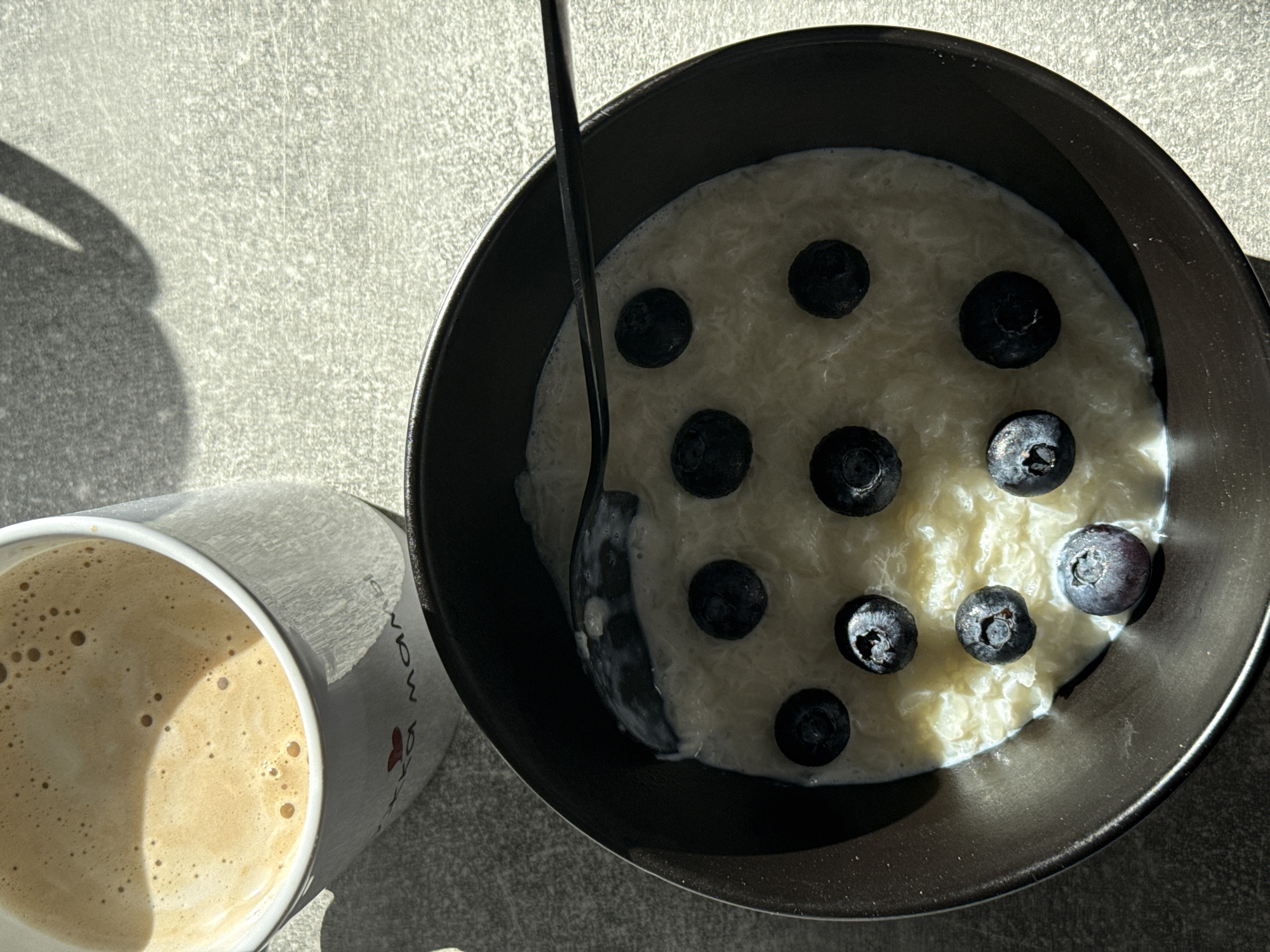 A bowl of rice pudding topped with blueberries and a mug of coffee sit on a textured gray surface. The coffee, in a white mug with a red heart and writing, is topped with foamy milk. A dark spoon is placed in the bowl of pudding. The scene appears to be indoors, possibly in a kitchen or dining area.