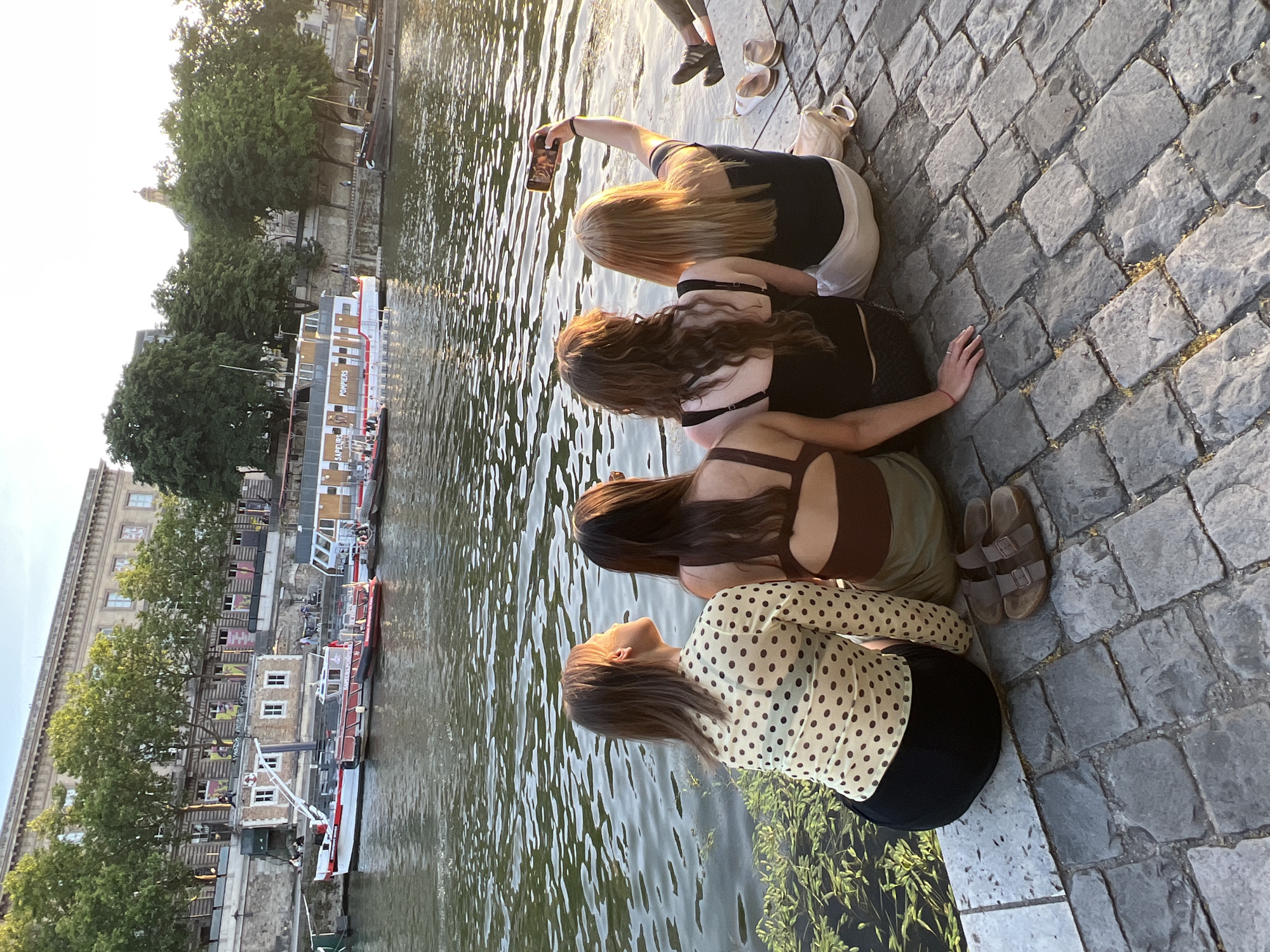 Four young women are sitting on a stone embankment beside a body of water, facing away from the camera. One of the women on the right is holding up a smartphone to take a selfie. The women are dressed casually in tops and skirts/pants. On the ground beside them are a pair of sandals. In the background, across the water, are several boats docked along the bank, and a large building with trees in front of it. This scene is likely along the Seine River in Paris, France. The lighting suggests it is late afternoon or early evening.