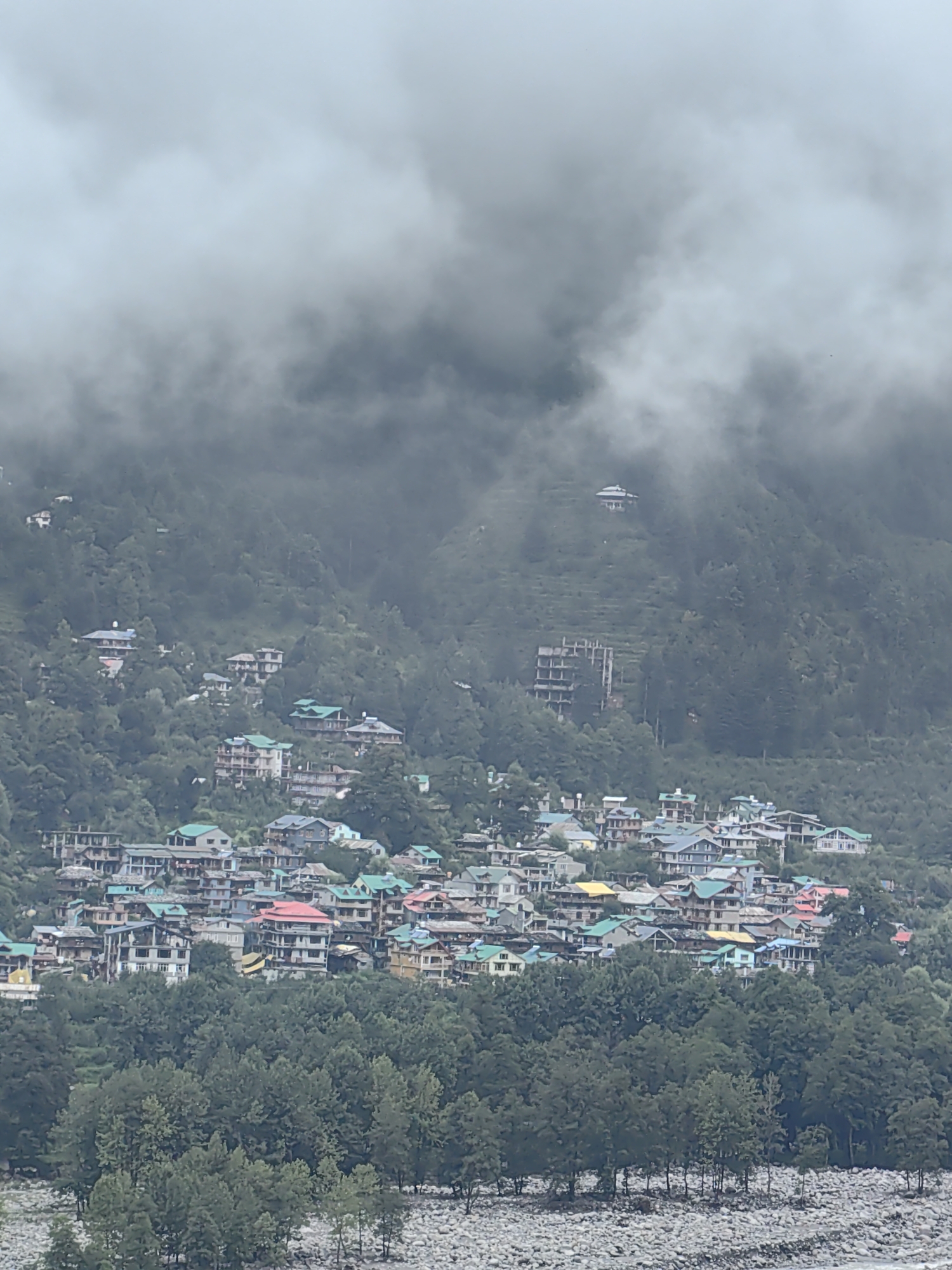 The image depicts a mountainous landscape with a town nestled on the slopes. The town, identified as Vashisht, India, is characterized by a dense cluster of buildings with colorful roofs, primarily green, red, and blue, interspersed with gray and white structures. The buildings appear to be multi-story, with some exhibiting modern architectural styles and others more traditional.
The town is situated on a steep, verdant hillside covered with trees, many of which are tall and dark green. The upper portion of the image is obscured by thick, low-lying clouds or mist, creating a hazy and atmospheric effect that partially veils the higher parts of the mountain and some of the distant buildings.
In the foreground, a rocky, barren area with scattered trees is visible, possibly a riverbed or a rocky clearing. The overall impression is one of a remote, mountainous settlement shrouded in mist. There are no people or animals visible in the image. The scene is captured from an elevated viewpoint, looking down upon the town and the surrounding natural environment.