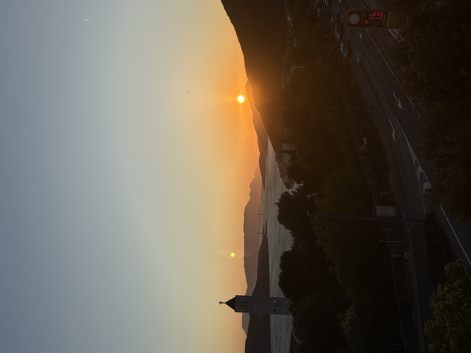 The image captures a sunset view in Neum, Bosnia and Herzegovina. The sun is positioned behind a mountain range, casting an orange glow across the sky and the water. A church tower is visible in the foreground, and a bridge stretches across the water in the midground. On the right side of the image, a road with cars and a gas station sign are visible.