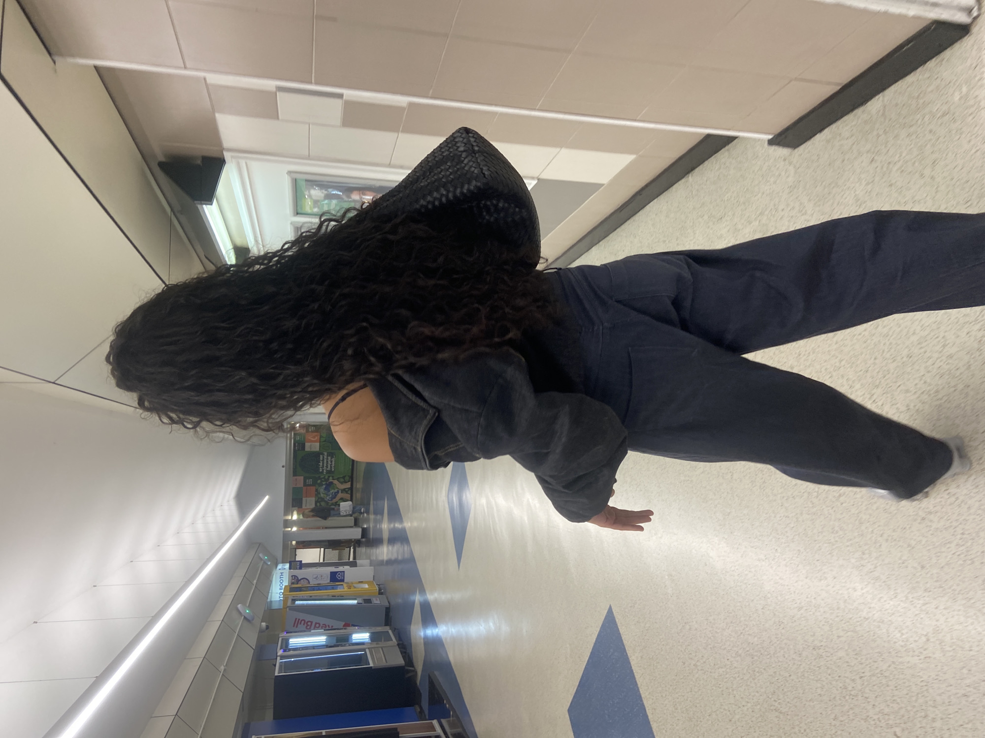 The image, oriented correctly, depicts a person from behind, standing or walking in a brightly lit indoor hallway. The person has long, dark, curly hair and is dressed in a dark-colored jacket, dark trousers, and a dark top, with a strap visible on the left shoulder. A black, woven-textured bag is carried over their left shoulder.
The setting is a modern public interior space, likely a corridor within a large building, shopping center, or facility in London, United Kingdom. The floor is predominantly light-colored with distinctive blue geometric patterns, resembling large arrows or triangles pointing along the hallway. The walls are light, and the ceiling features a series of integrated, long, bright LED strip lights, illuminating the space uniformly.
In the background, along the corridor, several commercial or informational displays are visible. One prominent display clearly shows the brand "Red Bull", and another sign reads "TO BOOTH". Further back, a large green-themed display contains text, though mostly unreadable, suggesting an informational or promotional nature, possibly related to environmental topics. The overall scene suggests a contemporary, functional walkway.