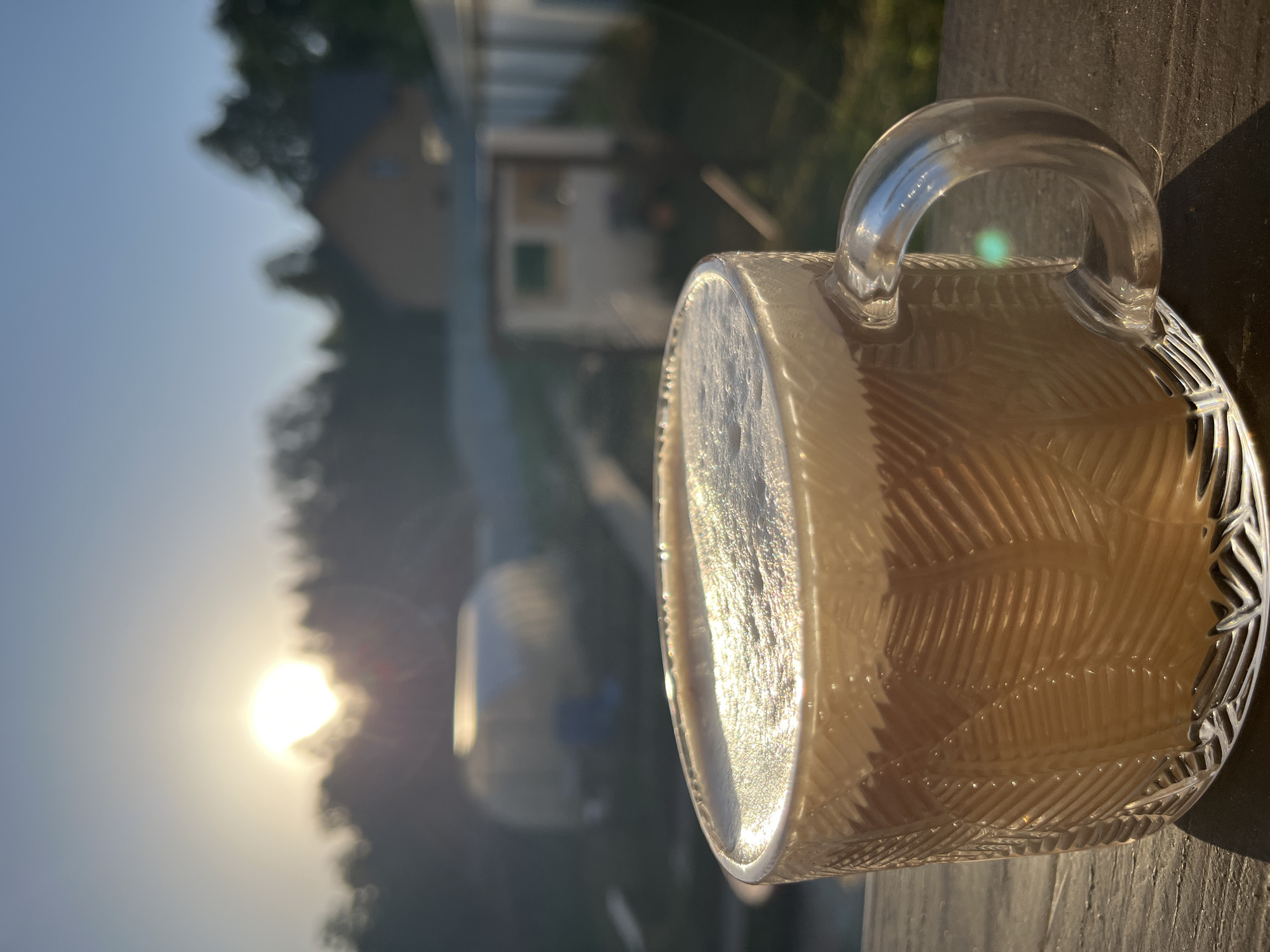 A close-up shot shows a clear glass mug with a decorative textured surface, filled with a light-colored beverage, likely coffee or tea with milk. The mug is resting on a weathered wooden surface, possibly a deck or windowsill. The background is blurred, but reveals a natural landscape with trees, a building with a light-colored exterior, and a bright sun positioned low in the sky, suggesting either sunrise or sunset. The location in the city of Chaikovsky, Russia, cannot be definitively determined from the image alone. The setting suggests an outdoor location with a residential area in the distance.