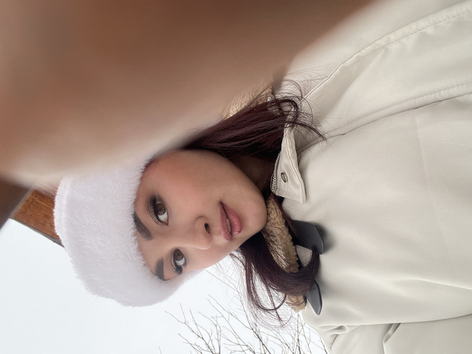 A young adult female is depicted in a close-up, upward-angled selfie, with her gaze directed towards the camera. She has fair skin, dark eyes, dark eyebrows, and dark red hair. Her lips are slightly parted, revealing a light pink lip gloss. She is wearing a white, fluffy, fur-trimmed hat and a light beige winter jacket with a brown faux fur collar lining. Two black, strap-like elements are visible near the collar of her jacket. A portion of a finger or thumb from the camera-holding hand obscures the upper right section of the image. The background consists of an overcast, light grey sky and bare tree branches to the left. A dark wooden architectural element is partially visible in the upper left background. The scene is outdoors in Neftekamsk, Russia.