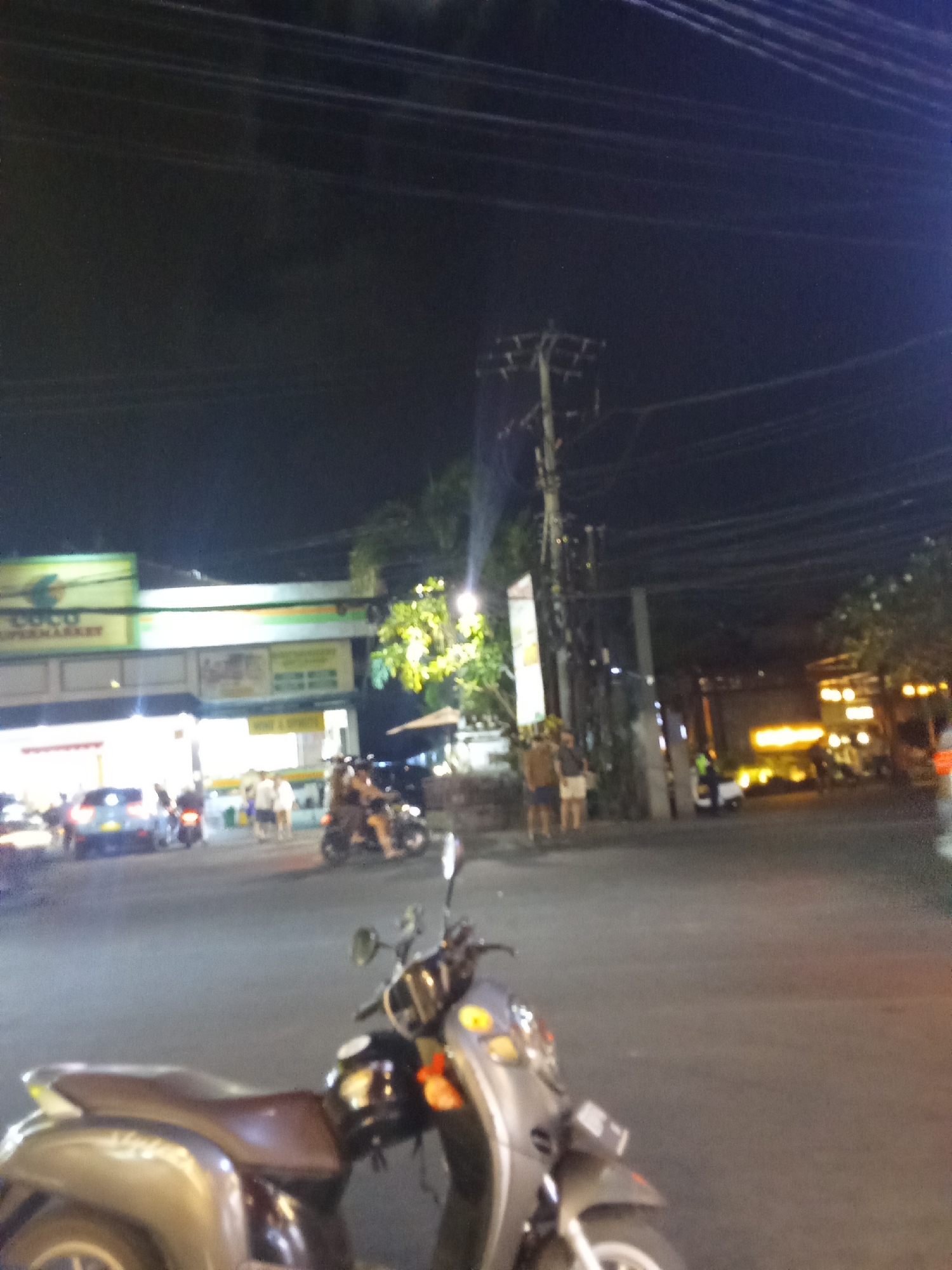 A nighttime, slightly blurry, street scene in Ubud District, Indonesia, captures a busy road with several vehicles and pedestrians. In the foreground, a silver scooter is parked. The background shows a supermarket with bright signage, a street with traffic including cars and other scooters, and several people walking and standing. Trees and utility poles are also visible, with overhead power lines contributing to the urban atmosphere. The lighting suggests it is after sunset, with artificial lights from buildings and vehicles illuminating the scene.
