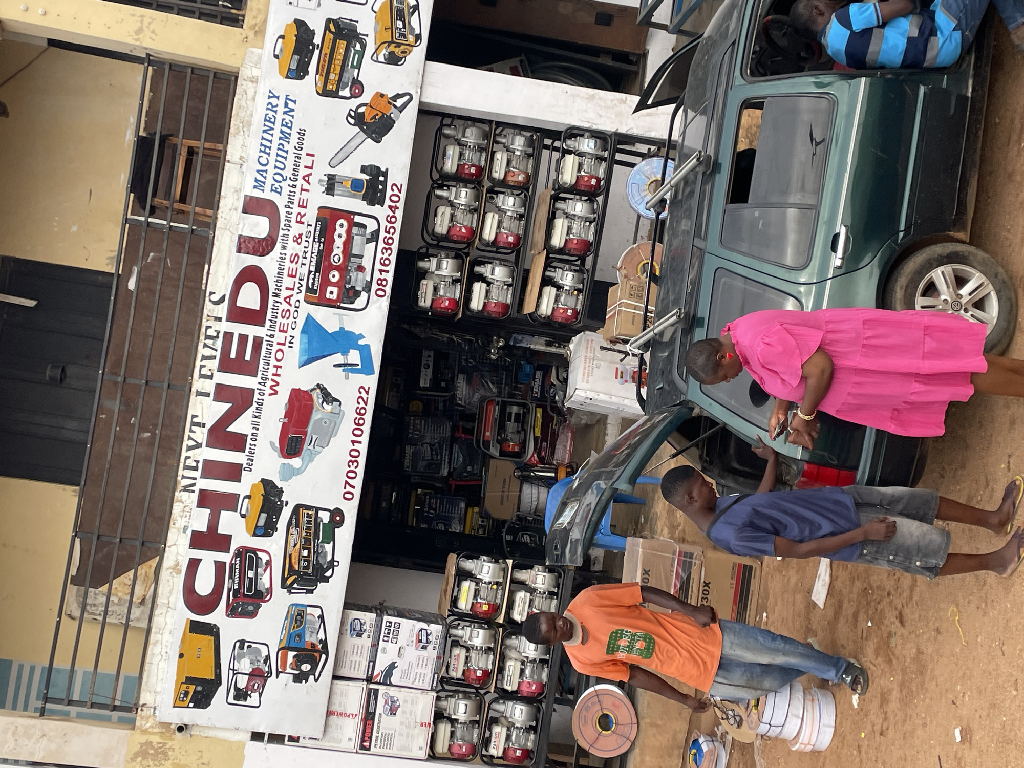 The photo depicts the storefront of a business named "CHINEDU" in Jos, Nigeria. The store sells "all kinds of Agricultural & Industry Machineries with Spare Parts & General Goods, WHOLESALES & RETALI (Retail)". The sign displays images of generators, chainsaws, and other machinery. Customers are shown interacting with personnel near an open vehicle trunk outside the store. Various boxes and equipment related to the business are stacked nearby.