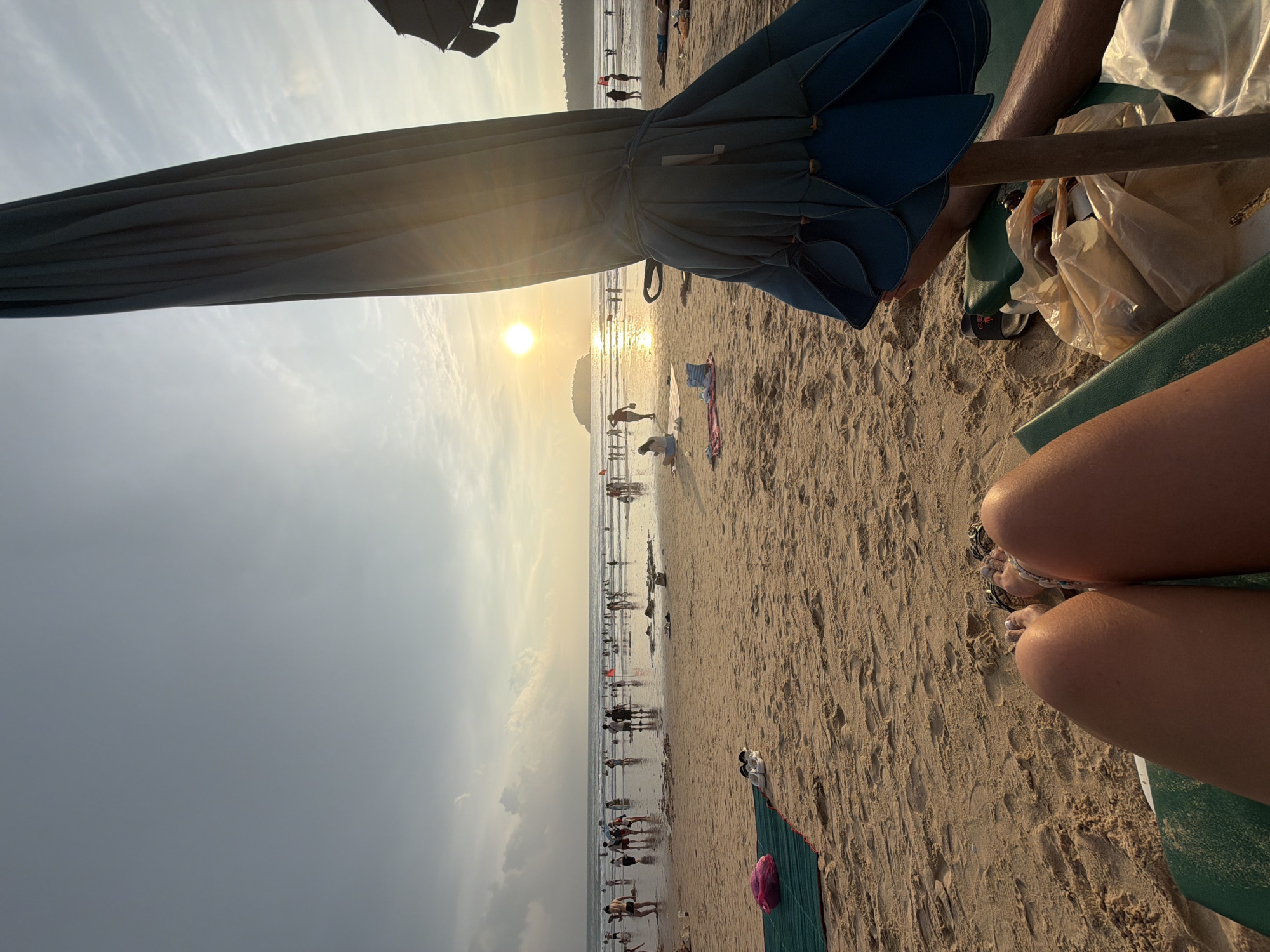 This is a first-person perspective shot of a beach in Karon, Thailand, at sunset. The foreground shows the legs and feet of a person sitting on a lounge chair, with a closed blue beach umbrella to their right. The sand is golden and textured, with footprints visible. In the middle ground, a shallow body of water reflects the setting sun, which is a bright orange orb low on the horizon. Numerous people are scattered across the beach and wading in the water, many of them silhouetted against the bright sky. There are a few small islands or landmasses visible in the distance. Red flags can be seen planted in the sand in several locations. The sky is a gradient of pale yellow near the horizon, transitioning to a grayish blue with scattered clouds.