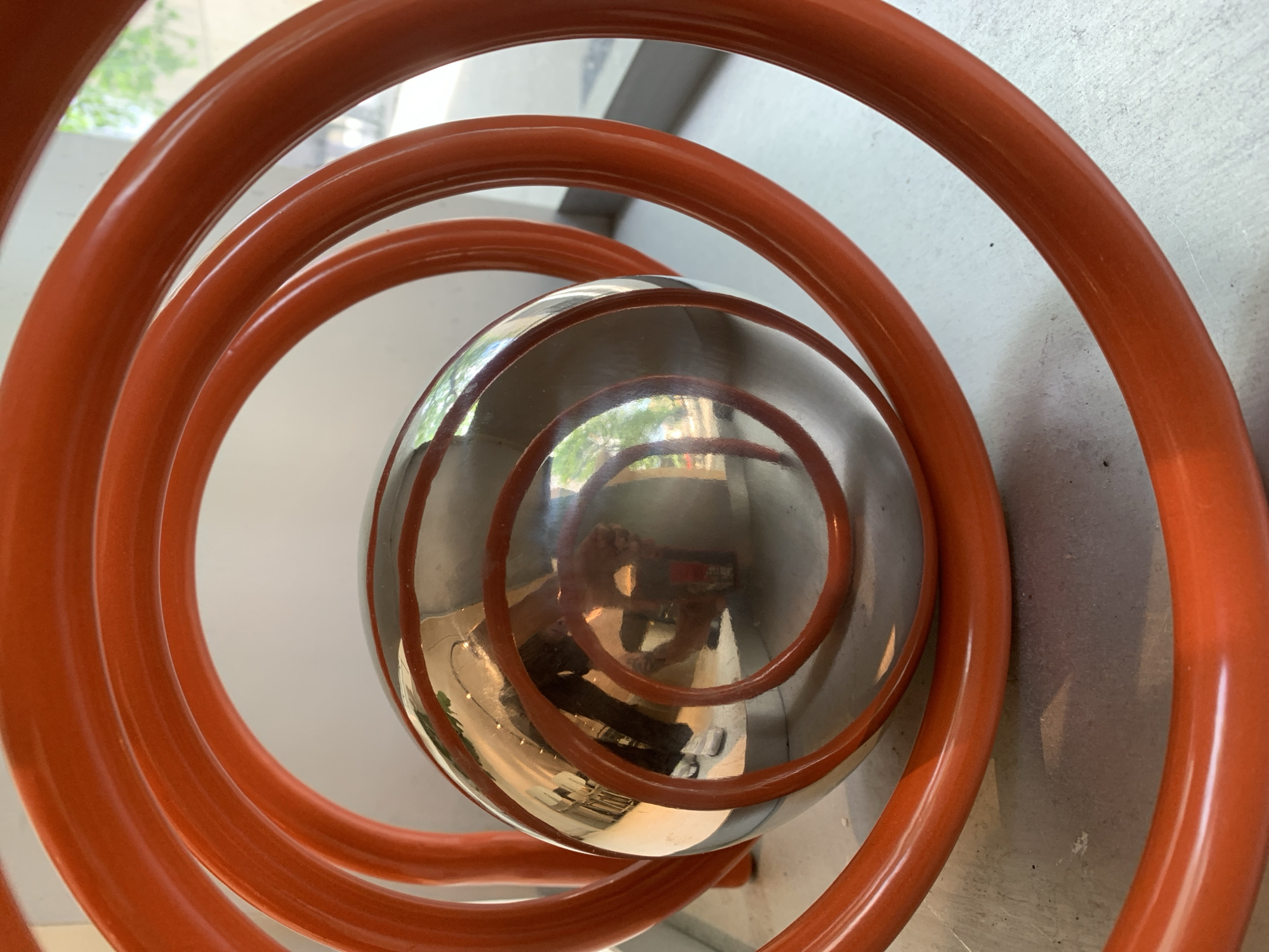 This image shows a close-up of a reflective sphere surrounded by a spiral of orange tubing. The sphere reflects the surroundings, including a person holding a phone taking the picture, portions of a building, and some greenery. The orange tubing forms a spiral pattern around the sphere, appearing to be part of the structure on which the sphere is placed. The background surfaces are light-colored and somewhat textured. The image is likely taken in a public space. Based on the design, architecture, and available public knowledge, this is likely a photograph of the Antoni Tàpies Foundation in Barcelona, Spain, specifically the "Cloud and Chair" sculpture on the roof. The sphere acts as a reflective surface capturing the surrounding environment.