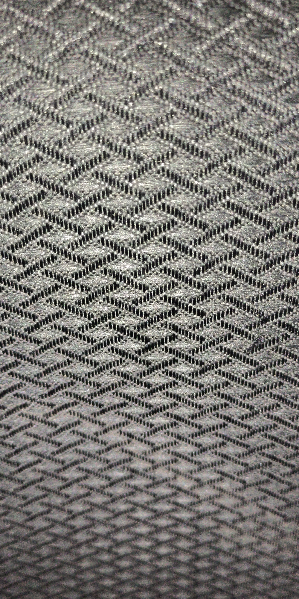 A close-up, top-down, full shot shows a black and white textured fabric with a diamond pattern. The fabric has a woven appearance with small holes creating the diamond shapes. The lighting creates a slight shadow effect on the bottom of the image, suggesting the fabric is draped or lying flat. There are no people or identifiable objects other than the fabric. The location is unknown.