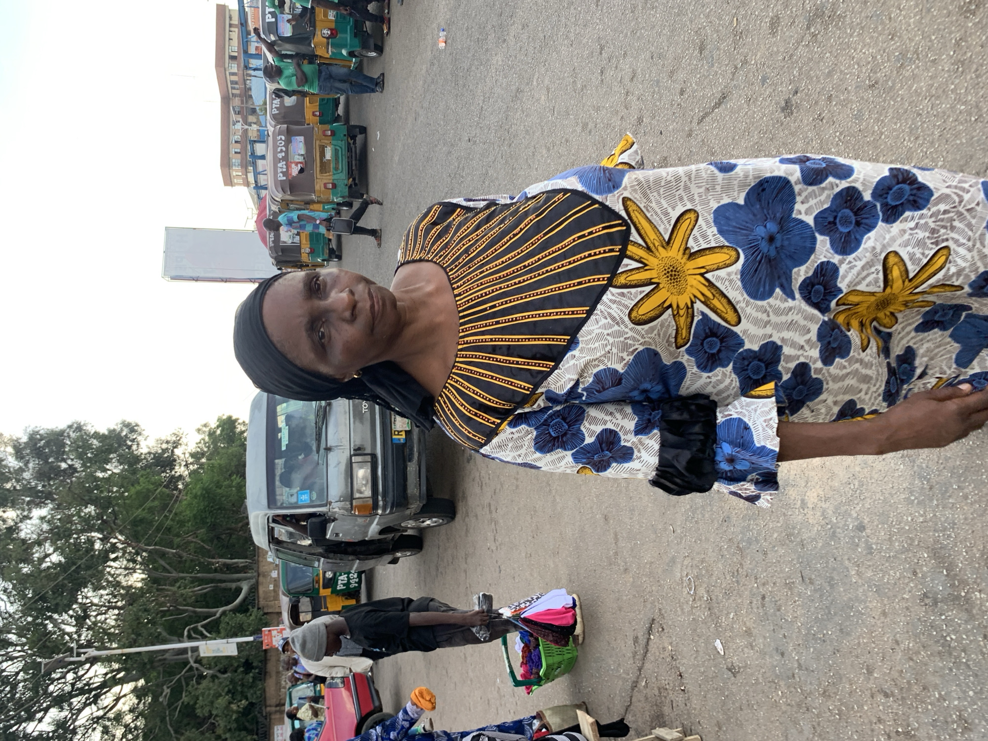 A medium-full shot captures a woman in the foreground, standing on a paved surface, likely a street or market area. She is wearing a black headscarf and a colorful, patterned dress with blue flowers and yellow starburst designs. The dress has a black yoke with yellow stripe accents. Her expression is neutral as she looks towards the camera.
Behind her, the scene depicts a bustling urban environment, consistent with a transportation hub or market in Jos, Nigeria. Several motor vehicles are visible, including a grey minivan, multiple auto-rickshaws (known locally as "Keke Napep"), and what appears to be a red car. The auto-rickshaws are lined up, suggesting they are either waiting for passengers or parked. People are scattered throughout the background, some walking, others seemingly engaged in commerce or awaiting transportation. The ground is paved with asphalt, and there are some trees and buildings visible in the distance, characteristic of an urban landscape. The lighting suggests it is either early morning or late afternoon. A green basket filled with assorted colorful items, possibly goods for sale, is situated to the left of the woman, held by a young person.