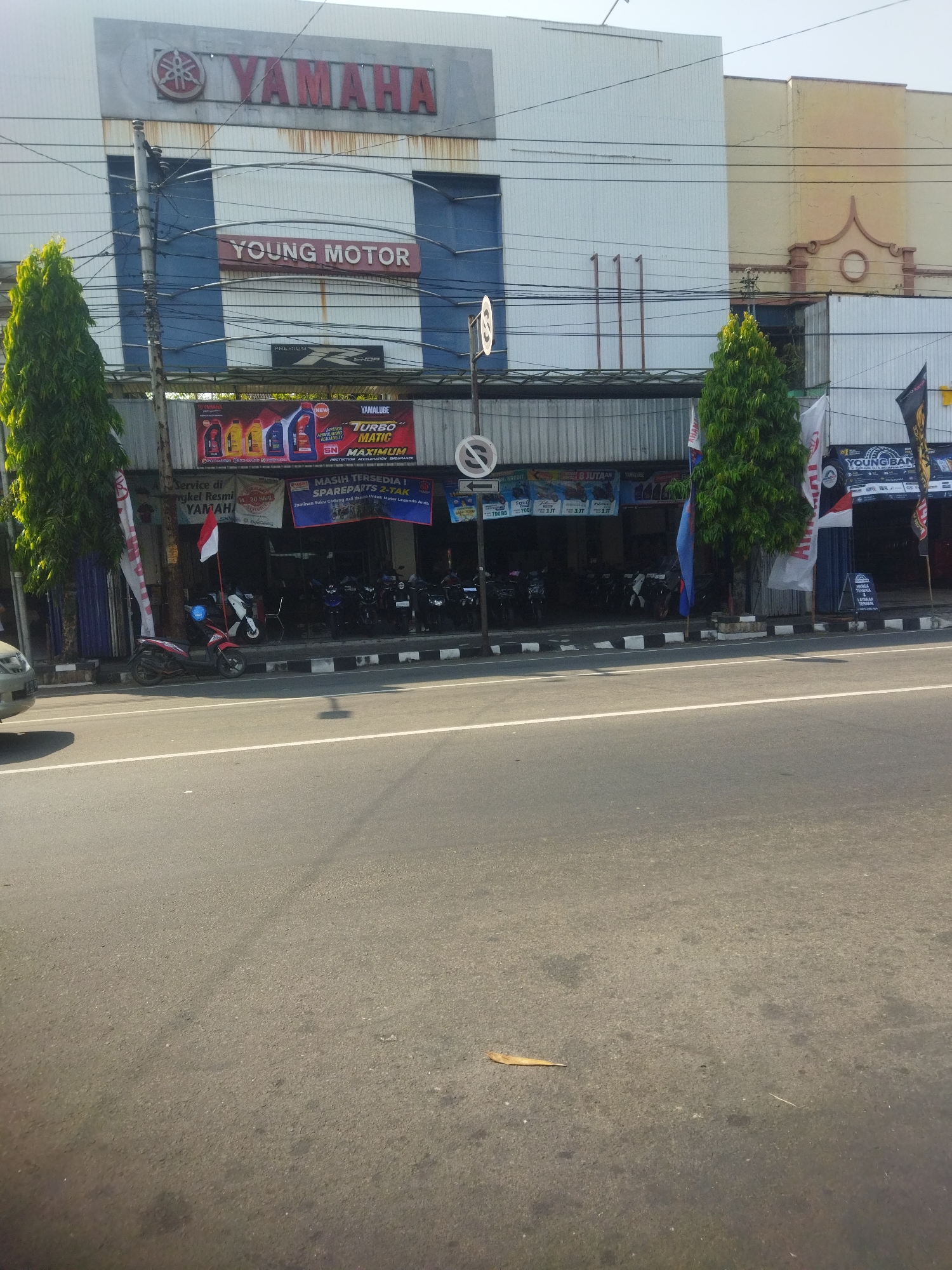 The image depicts the exterior of a Yamaha dealership called "Young Motor" in Giripurwo, Indonesia. The building has a light-colored facade with blue trim. A large "YAMAHA" logo is prominently displayed above the entrance. Below this, "YOUNG MOTOR" is written in red letters on a maroon background.
Several motorcycles are parked in front of the establishment, some are displayed for sale. A red and white Indonesian flag is mounted on a pole next to a motorcycle. Banners and advertisements for Yamaha products, including engine oil and spare parts, are visible. Two tall, green trees stand on either side of the building's entrance area.
The street in front of the dealership is paved and has white lane markings. A silver car is partially visible on the left side of the image, parked on the side of the road. The scene appears to be outdoors during daylight, with a clear sky. Electrical wires are strung across the building and overhead.