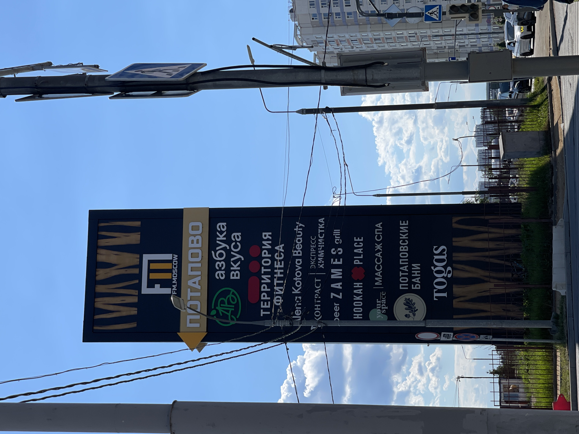 The image captures an outdoor urban or suburban scene, dominated by a large, vertical advertising pylon under a clear blue sky with scattered white cumulus clouds. The time of day is bright daylight, suggesting a sunny day.
The primary subject is the black and yellow signpost, which lists various businesses in Russian and English. Visible text includes:
* "FM.MOSCOW"
* "ПОТАПОВО" (Potapovo)
* "азбука вкуса" (azbuka vkusa - "Alphabet of Taste," a grocery chain)
* "ТЕРРИТОРИЯ ФИТНЕСА" (Territoria Fitnesa - "Fitness Territory")
* "lena Kotova Beauty"
* "КОНТРАСТ ЭКСПРЕСС ХИМЧИСТКА" (Kontrast Express Himchistka - "Contrast Express Dry Cleaning")
* "Deer ZAMES grill"
* "HOOKAH PLACE"
* "your space МАССАЖ СПА" (your space MASSAGE SPA)
* "ПОТАПОВСКИЕ БАНИ" (Potapovskie Bani - "Potapovo Baths")
* "Togas"
Above the sign, a utility pole runs horizontally across the frame, supporting multiple wires and traffic signs, including a blue square pedestrian crossing sign. In the background, modern multi-story residential buildings are visible, confirming an urban setting. A paved road or sidewalk runs along the bottom, with a metal fence and some greenery separating it from further structures or undeveloped land. A few parked cars are faintly discernible in the distance. No specific human activity is taking place, but the commercial signage and infrastructure indicate a lively area in Moscow, Russia.