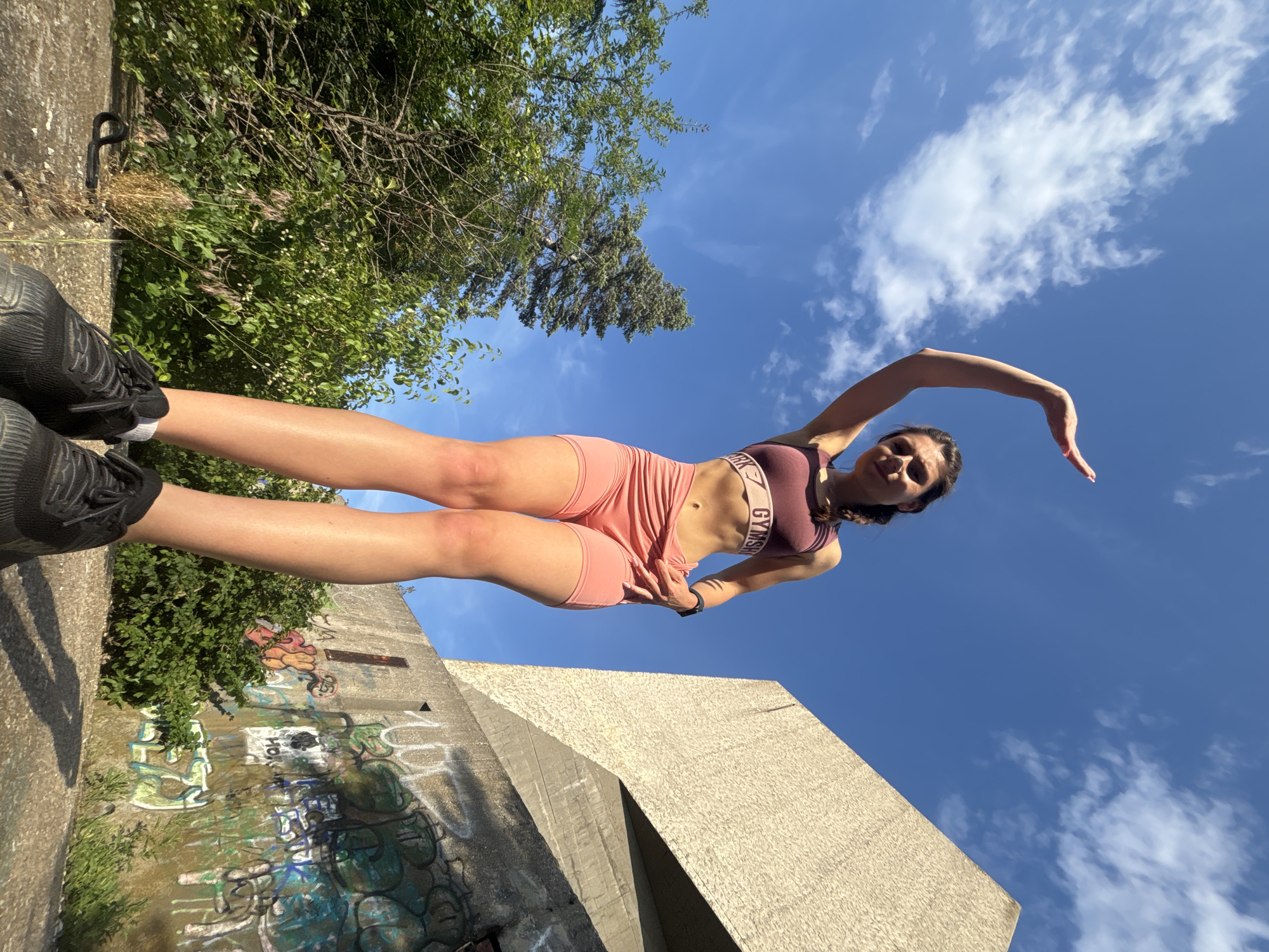 A young woman with dark hair braided down her back is standing outdoors, looking up at the sky with her right arm raised above her head. She is wearing a maroon sports bra with "GYMSHARK" written across the band, and matching coral-colored shorts. Her black sneakers are visible at the bottom of the frame. The background shows a bright blue sky with scattered clouds, lush green trees, and a large, brutalist concrete structure covered in graffiti. The location is the northern part of the city of Varna, Bulgaria, specifically the area around the former "Balkantourist" sanatorium, an abandoned concrete building with distinctive architectural features. The lighting suggests it is daytime, likely late morning or early afternoon, given the strong sunlight.
