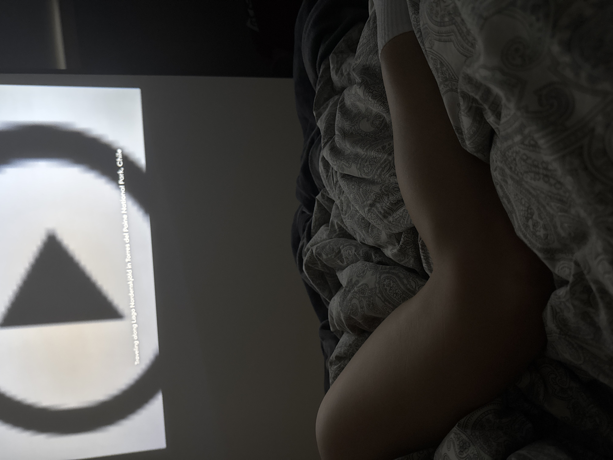 The image depicts a person lying in bed, covered by patterned bedding. Next to the bed, a projection screen displays a black-and-white image of a triangle enclosed within a circle. Text along the side of the projection reads "Traveling along Lago Nordenskjöld in Torres del Paine National Park, Chile." The location of the bed and the projection within a specific city or place is unknown.