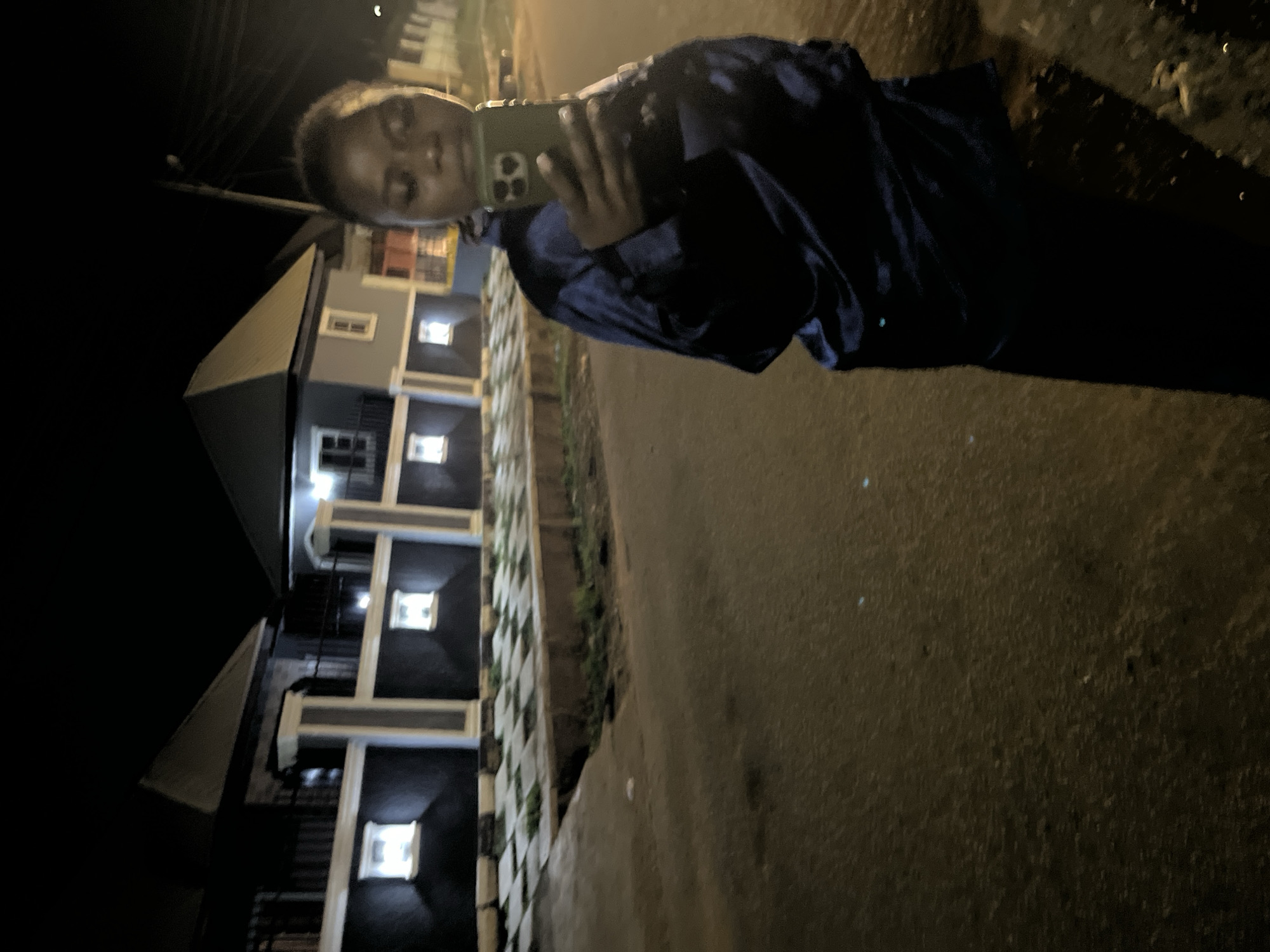 A full-body, nighttime, street-level selfie is captured in Abuja, Nigeria. A young woman, facing the camera, holds up a green smartphone in her right hand, taking the picture. She is wearing a shiny, dark blue, long-sleeved jacket. The background features modern, two-story houses with dark facades, lighter-colored trim, and illuminated windows. A checkered paved area separates the houses from the street, with some overgrown grass. The street itself is asphalt and appears wet, reflecting some of the ambient light. The overall scene is dimly lit, illuminated by artificial lights from the houses and possibly streetlights, creating long shadows.