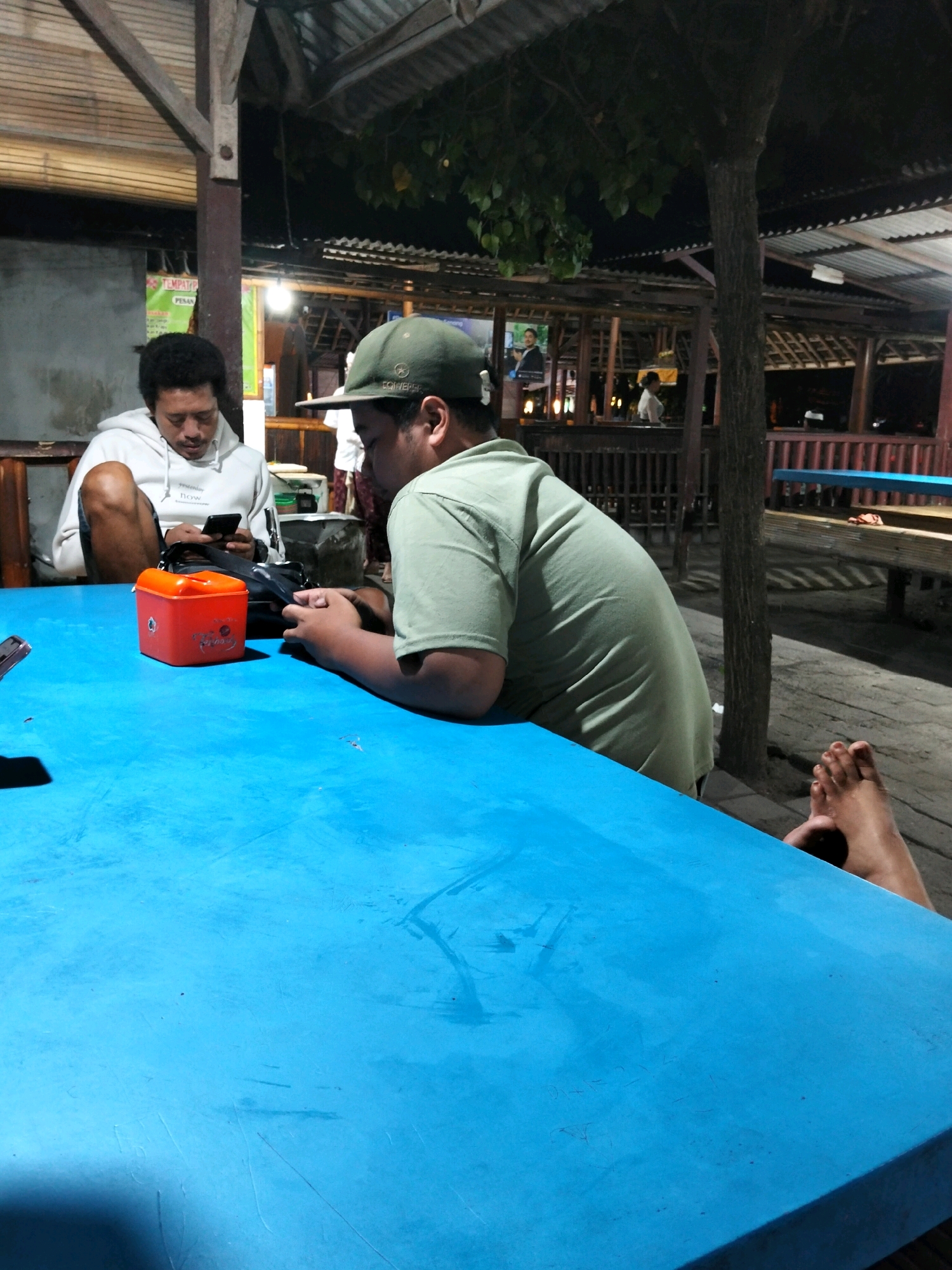 Two men are seated at a blue plastic table outdoors at night. The man on the left is wearing a white hooded sweatshirt and is looking down at a black smartphone in his hands. He has dark, curly hair. The man on the right is wearing a green t-shirt and a green baseball cap. He is leaning forward, with his hands on the table, looking down. An orange rectangular container with a black lid is placed on the table between the two men. The background shows a rustic outdoor seating area with wooden structures and roofing. Trees are visible, and there are some lights illuminating the area. One of the man's feet is visible resting on the ground to the right of the table. The location is likely a casual eatery or gathering spot.