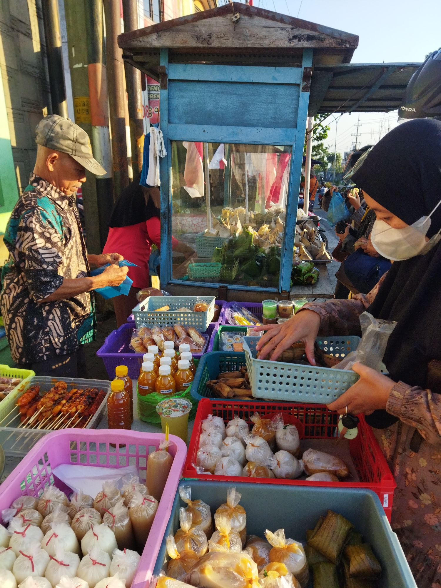 This is a medium shot, taken at eye level, of an outdoor food stall in Semarang, Indonesia. The stall is located on a street with other stalls and pedestrians visible in the background. The stall has a blue wooden structure with a metal roof. A man wearing a cap and a patterned shirt is standing on the left, preparing a blue plastic bag. To his right, a woman wearing a black hijab and a face mask is examining food items in a blue basket. In front of the stall, there are several baskets filled with various food items, including grilled skewers, bottled drinks, and wrapped snacks. The lighting suggests it is either early morning or late afternoon, with the sun casting long shadows. The overall scene depicts a typical street food vendor in an urban setting.