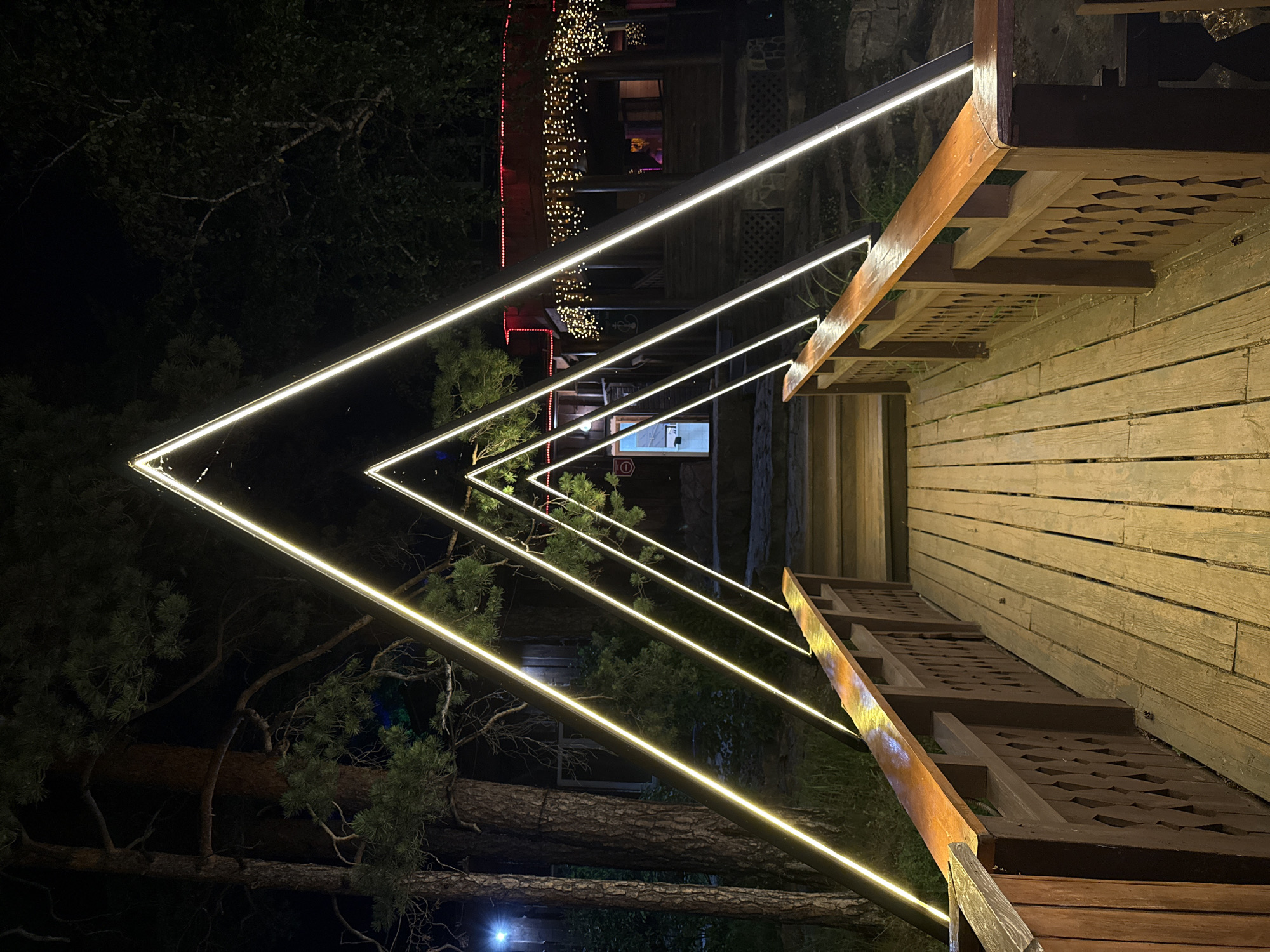 A perspective shot from a wooden walkway at night shows a series of three illuminated triangular arches. The walkway is made of weathered wooden planks and has wooden railings with decorative latticework. The arches are formed by bright, continuous yellow LED lights, receding into the background. Beyond the arches, a wooden building with a sloped roof is visible, adorned with strings of small, warm-toned lights. The building appears to be a gazebo or a covered outdoor seating area. Trees with dark foliage are present on the left and right sides of the scene. The overall environment suggests an outdoor recreational area, possibly a park or a resort, at Zolotoi Plyazh, Russia. No people are visible.