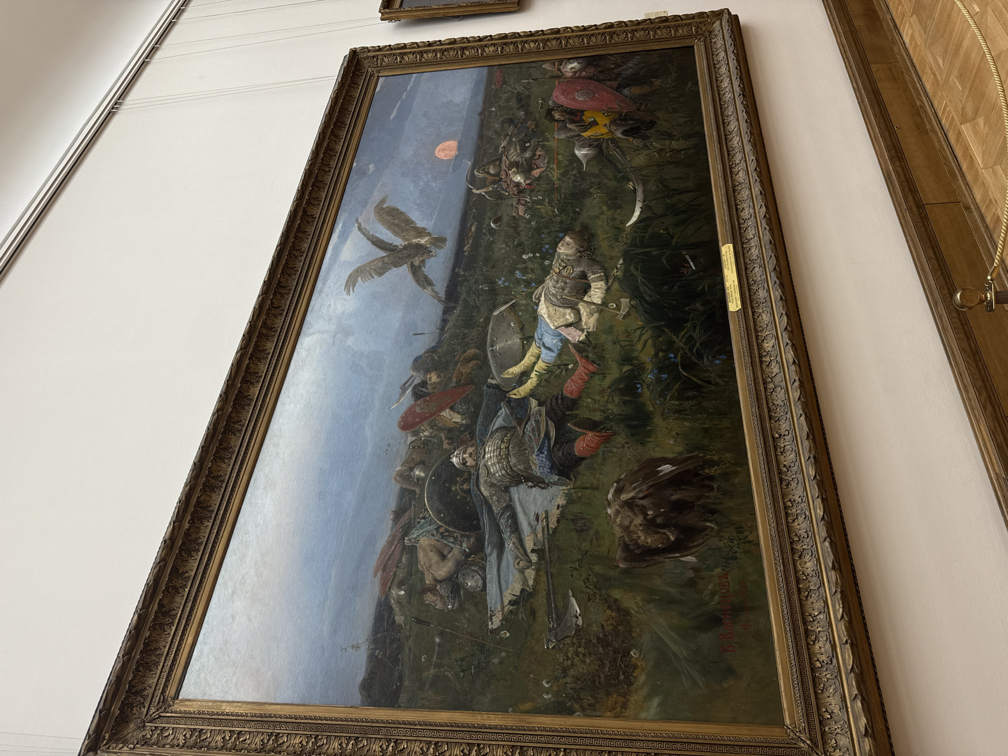 The image shows a painting in a highly ornate gold frame, displayed on a white wall. The painting appears to depict a battlefield scene with figures in armor, a large bird (possibly an eagle or vulture) flying overhead, and a red orb in the sky. The style appears classical, possibly romantic or historical. The painting is likely located in an art gallery in Moscow, Russia based on information about similar artworks found in Moscow.