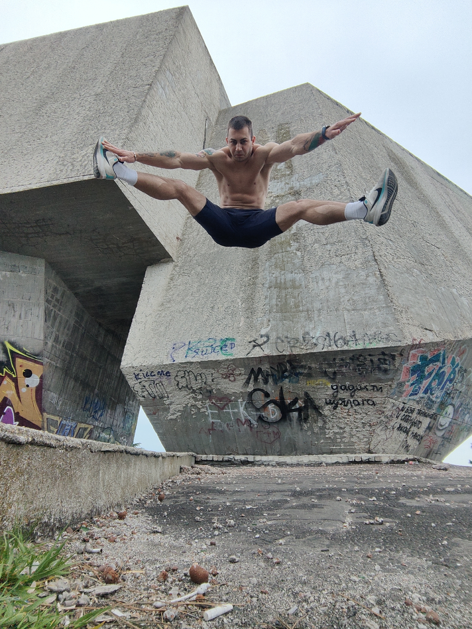 A shirtless, fair-skinned man is performing a split jump in front of a large concrete monument. He has short dark hair, dark shorts, white socks, and athletic shoes. His arms are extended to either side. The monument is covered in graffiti and appears to be in a state of disrepair. The ground below is littered with debris and small rocks. Green grass is visible in the lower left corner. The location is the Pantheon in Varna, Bulgaria.