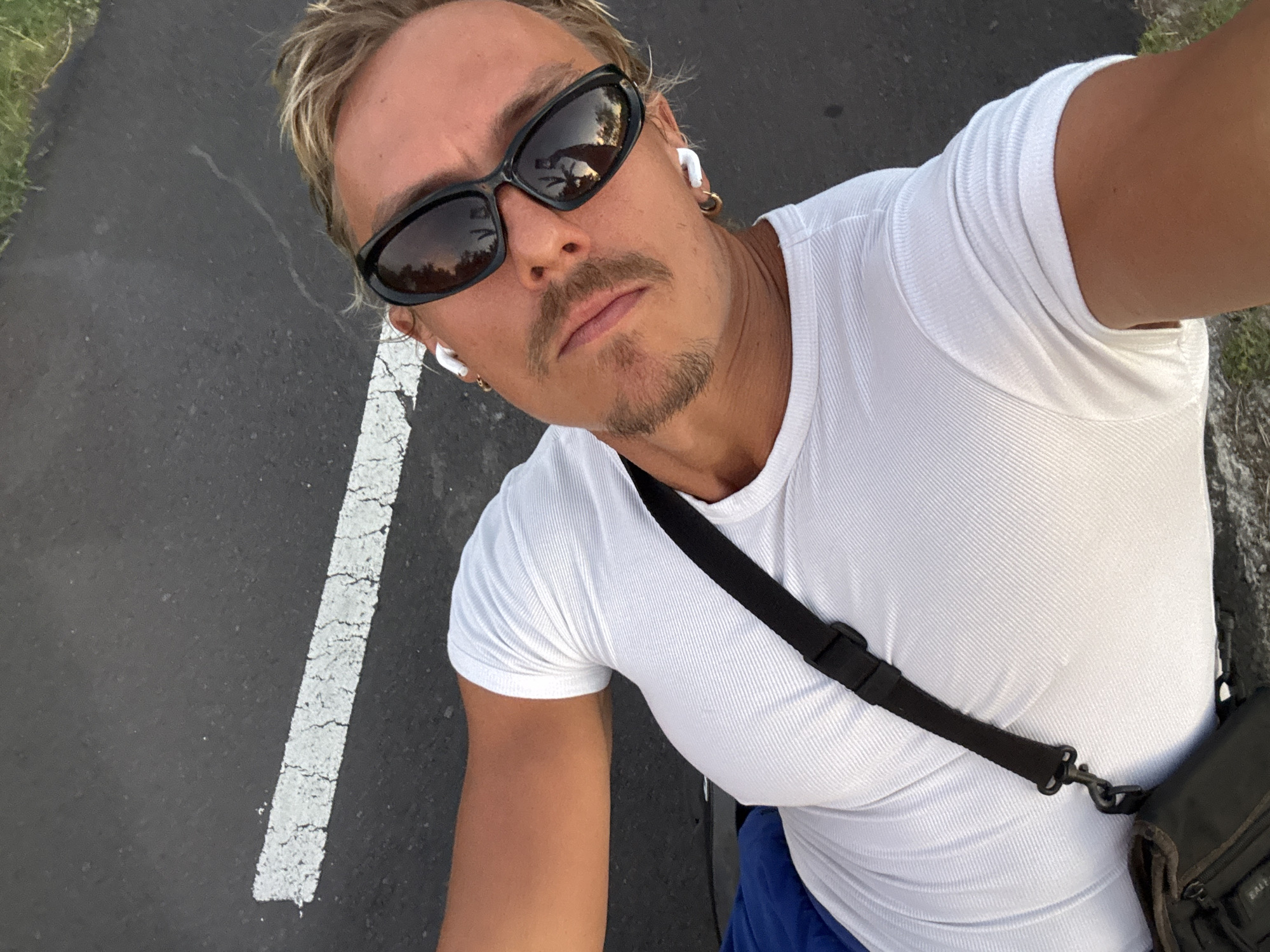 A man wearing sunglasses and a white shirt takes a selfie. He is on a paved road with a white line marking. The reflection on the sunglasses suggests an outdoor setting. The man has light brown hair with blond highlights and a mustache. He is wearing wireless headphones in his ears. He also has earrings on. The location cannot be determined to be in Selemadeg Timur, Indonesia from the image alone.